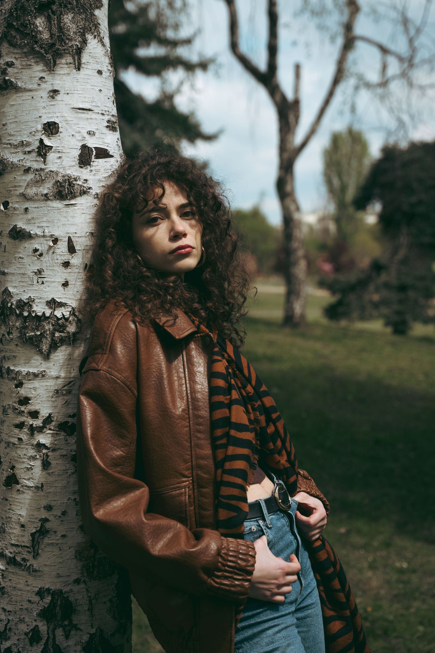 Curly-haired woman in brown leather jacket and tiger-print scarf leaning against birch tree in park.