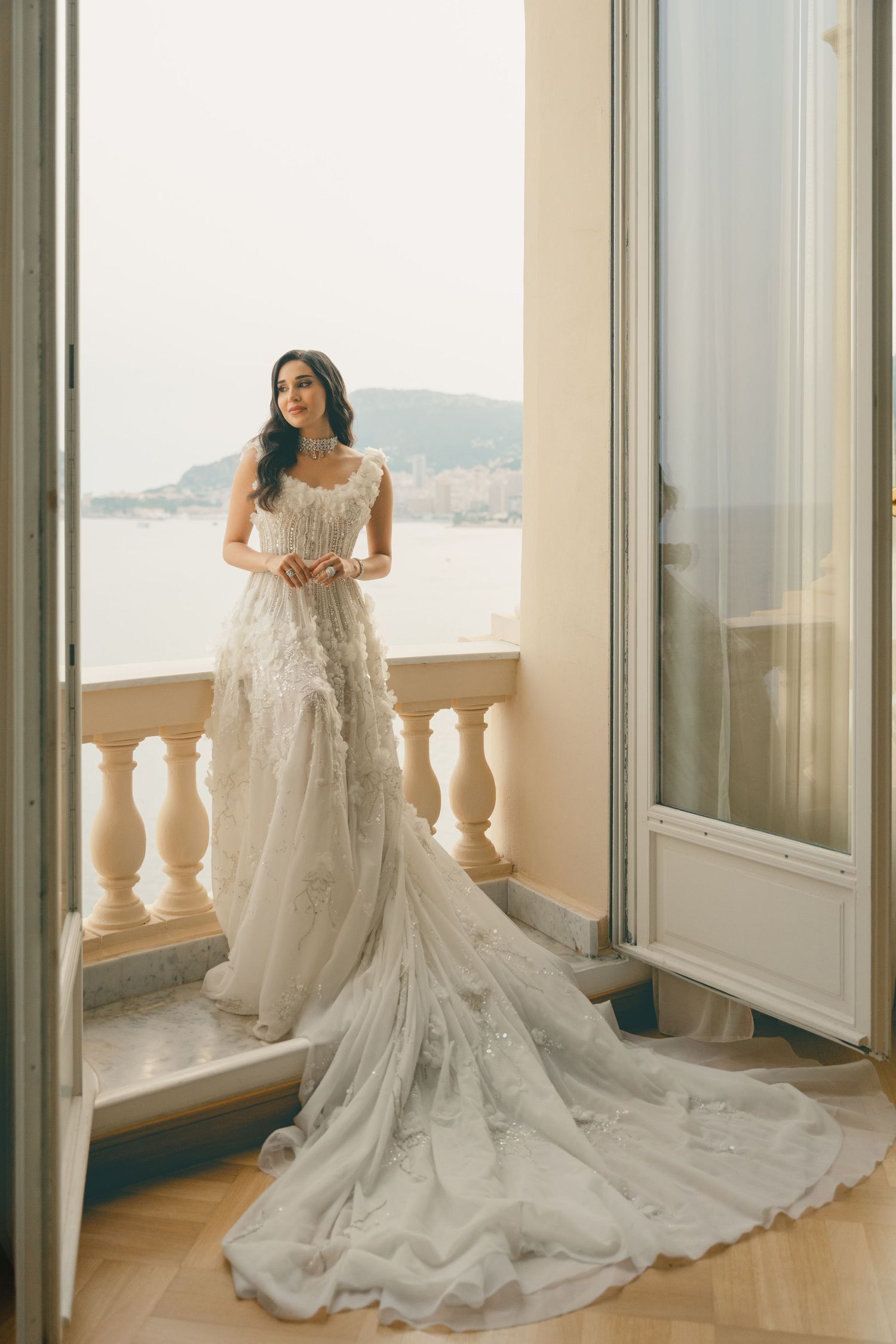 Bride in elegant white lace gown with long train stands on balcony overlooking scenic coastal view.
