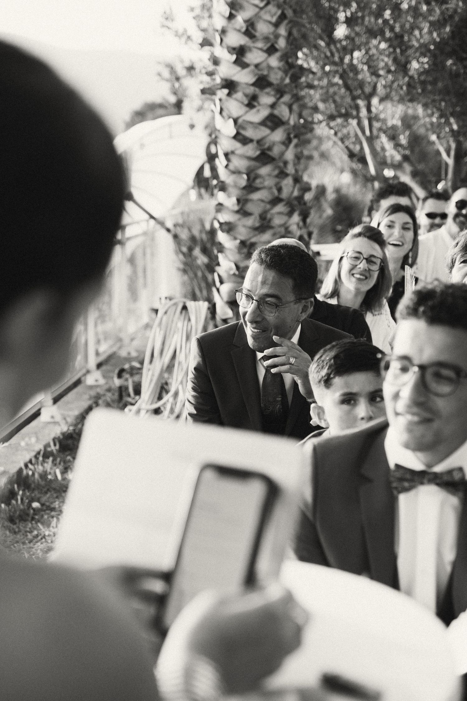 Black and white wedding ceremony photo of an emotional groom listening to vows among smiling guests.
