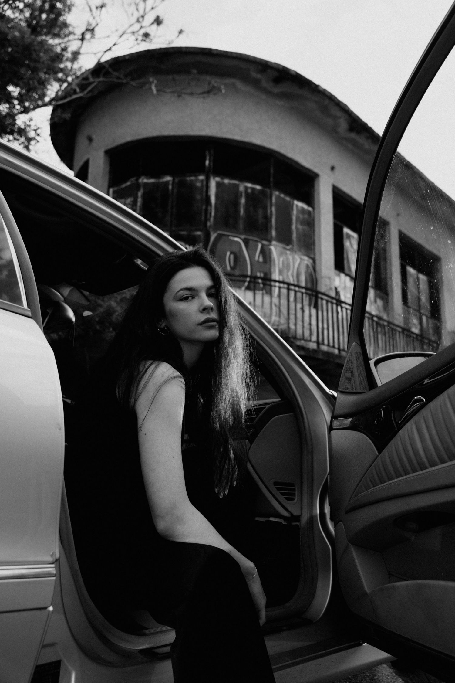 Black and white photo of woman exiting silver car near abandoned graffiti-covered building.
