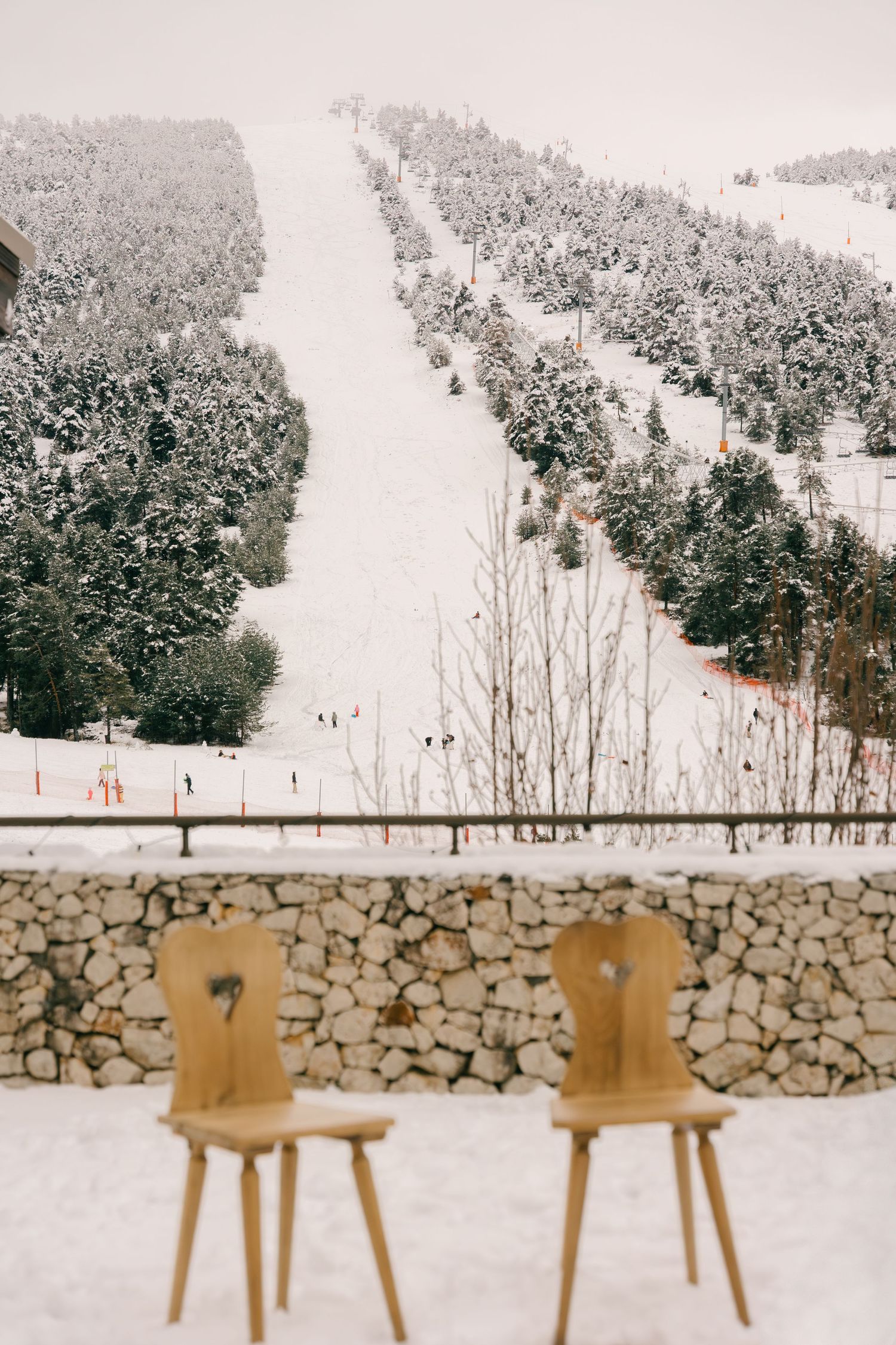 Two wooden chairs set before a stone wall overlooking a snowy ski slope with pine trees.