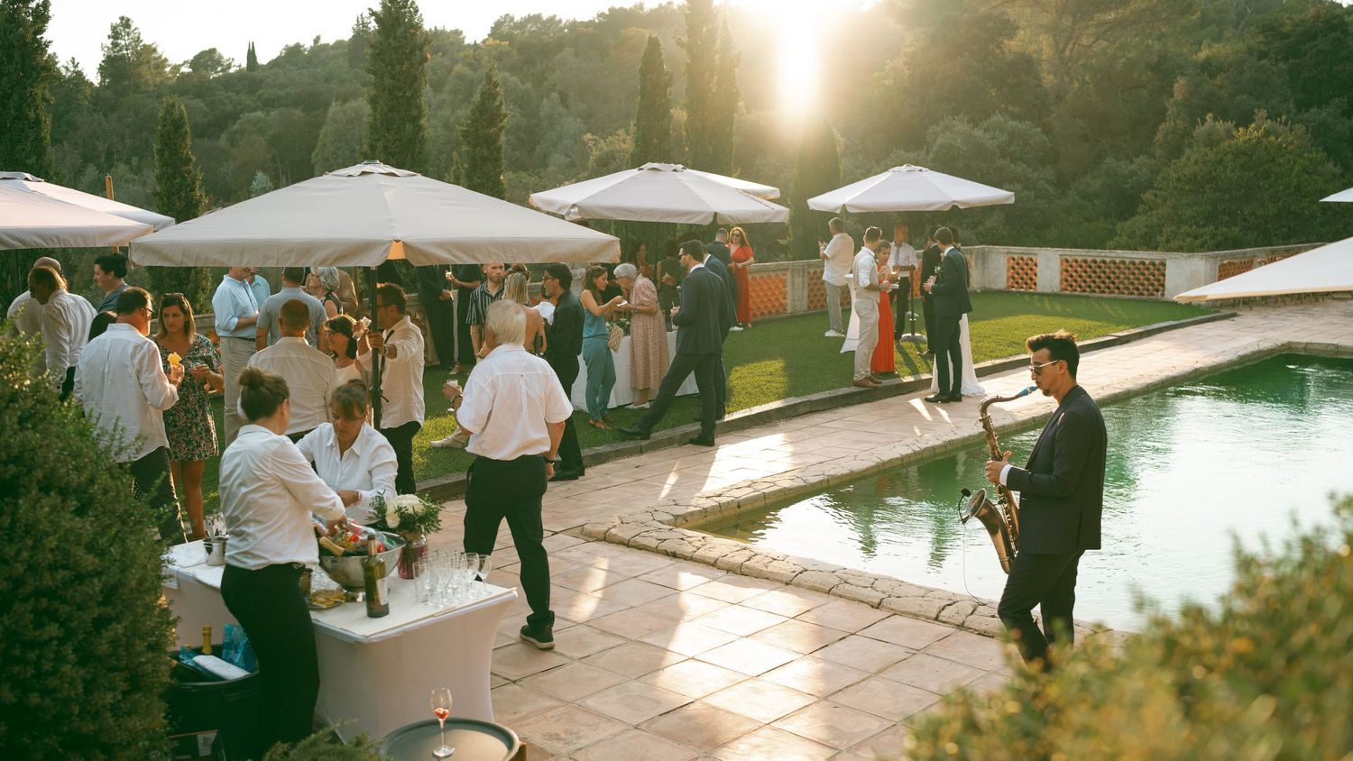 Elegant outdoor wedding reception by a pool with guests mingling, white umbrellas, and a saxophonist performing at sunset.