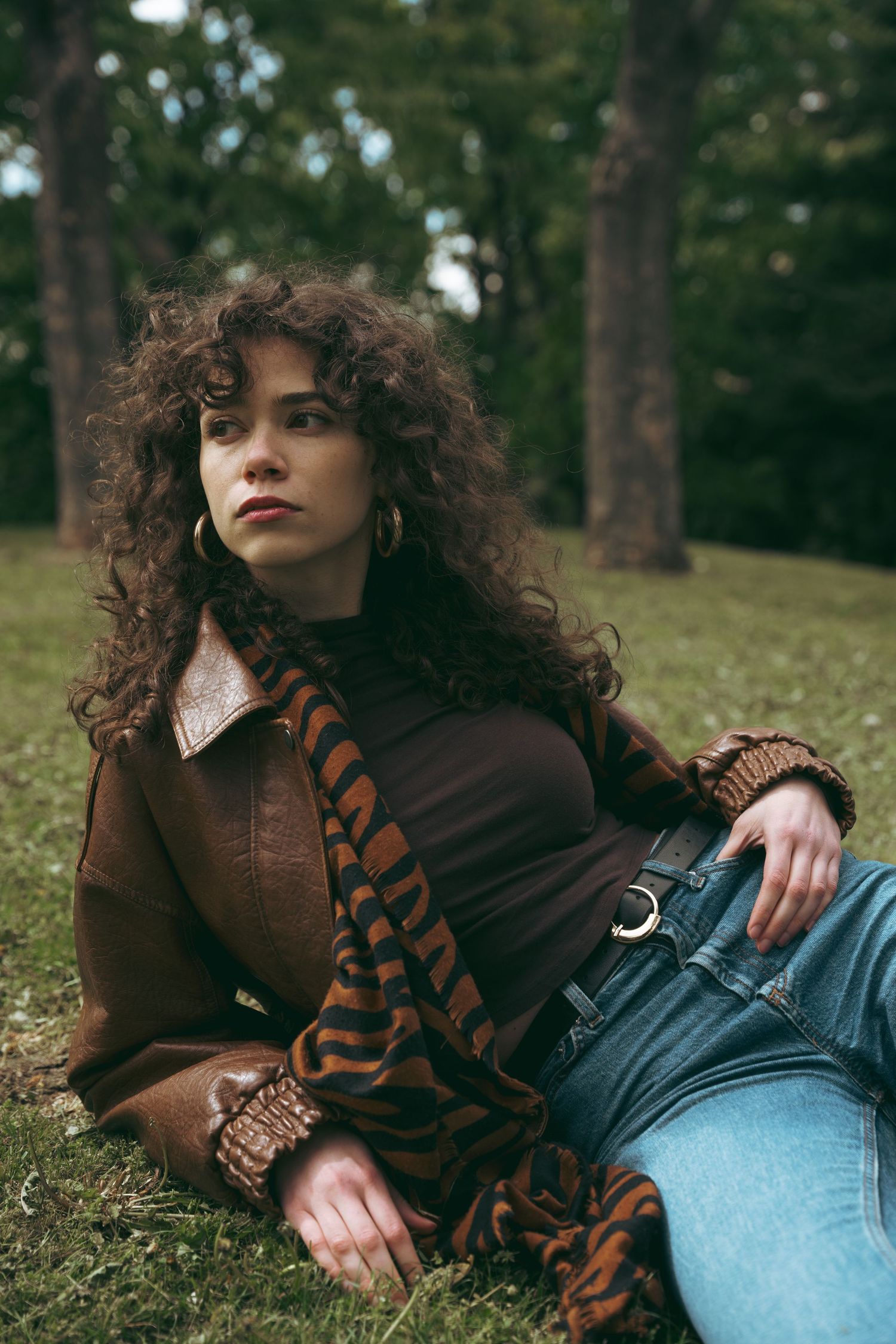 Stylish woman with curly hair in brown leather jacket and scarf reclining on grass in park.