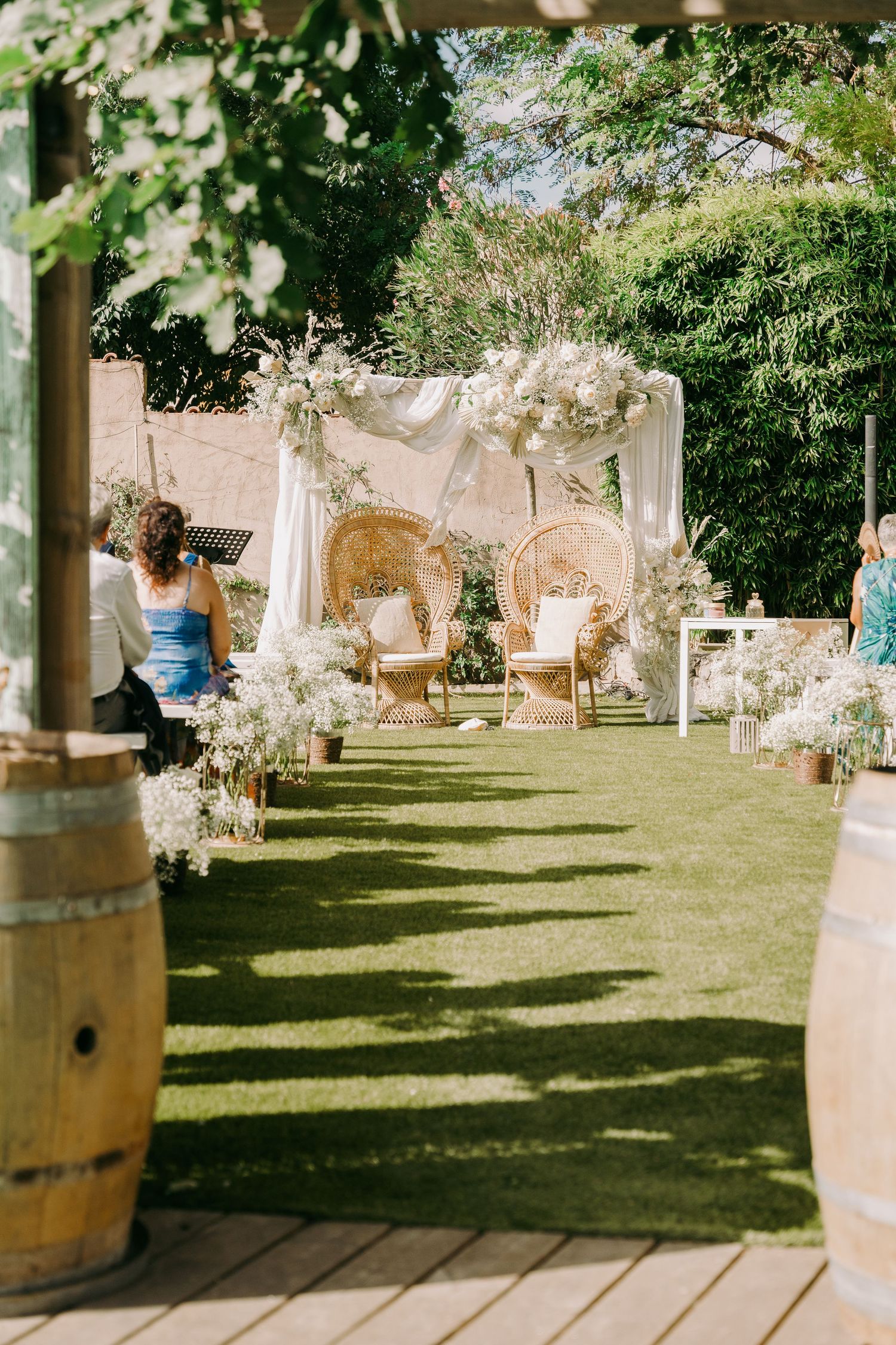 Elegant outdoor wedding ceremony setup with white floral arch, peacock chairs, and barrel decorations.