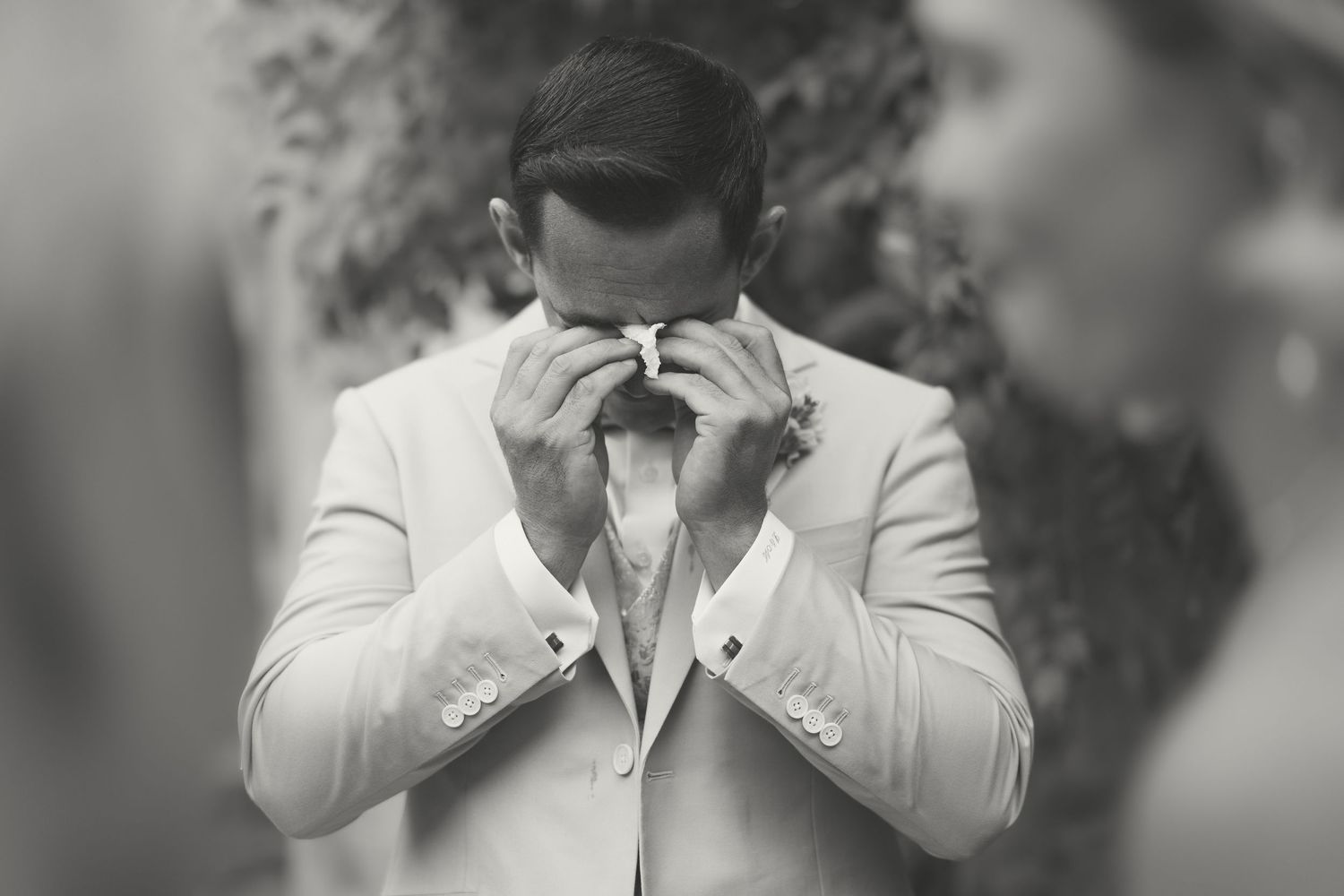 Emotional groom in white suit wiping tears during outdoor wedding ceremony, black and white photography.