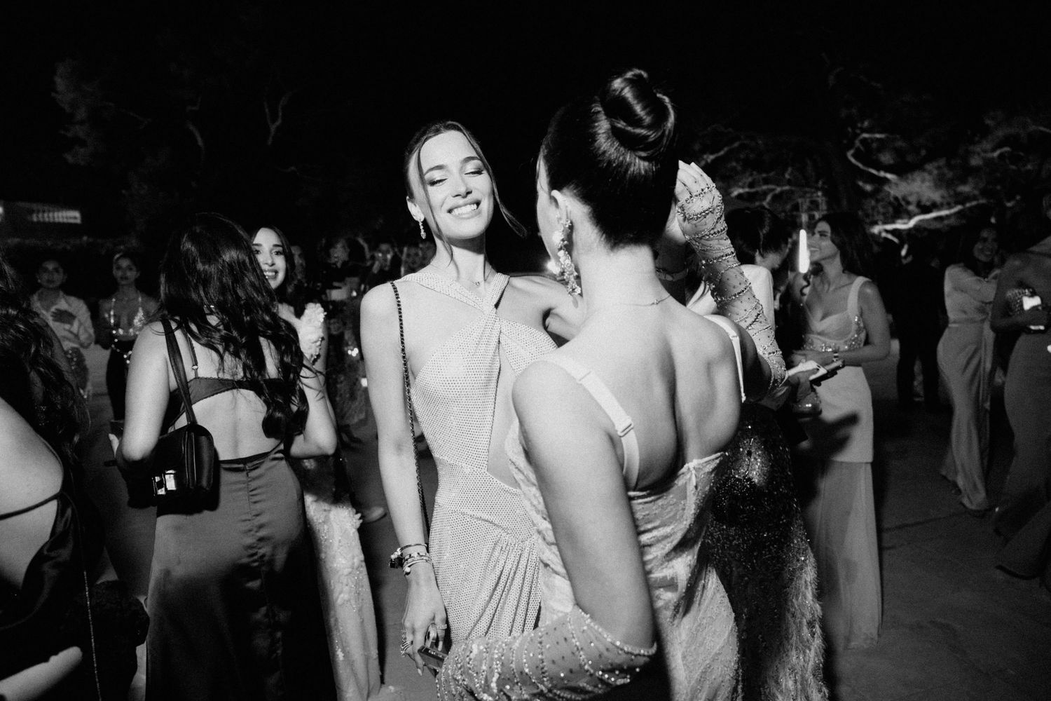 Two elegantly dressed women in gowns share a joyful moment at a glamorous evening party in black and white photo.
