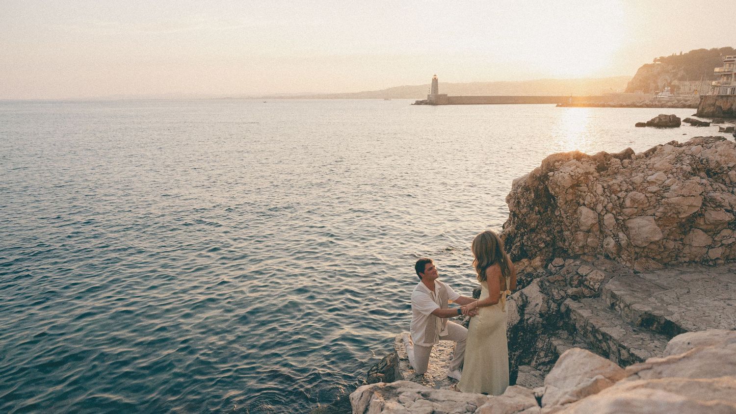Couple on rocky seaside cliff toasting champagne glasses during breathtaking golden sunset. Wedding Proposal
