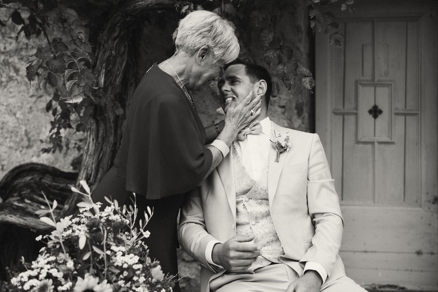 Black and white wedding photo of two brides sharing a tender moment, one touching the other's face outdoors.