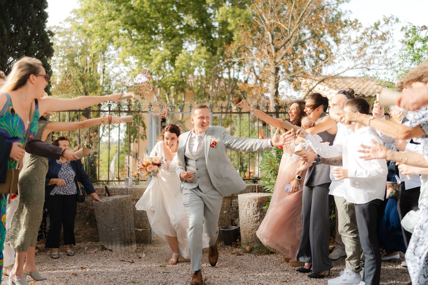 Joyful newlyweds run through a celebratory guest tunnel at an outdoor wedding ceremony in a sunny garden setting.
