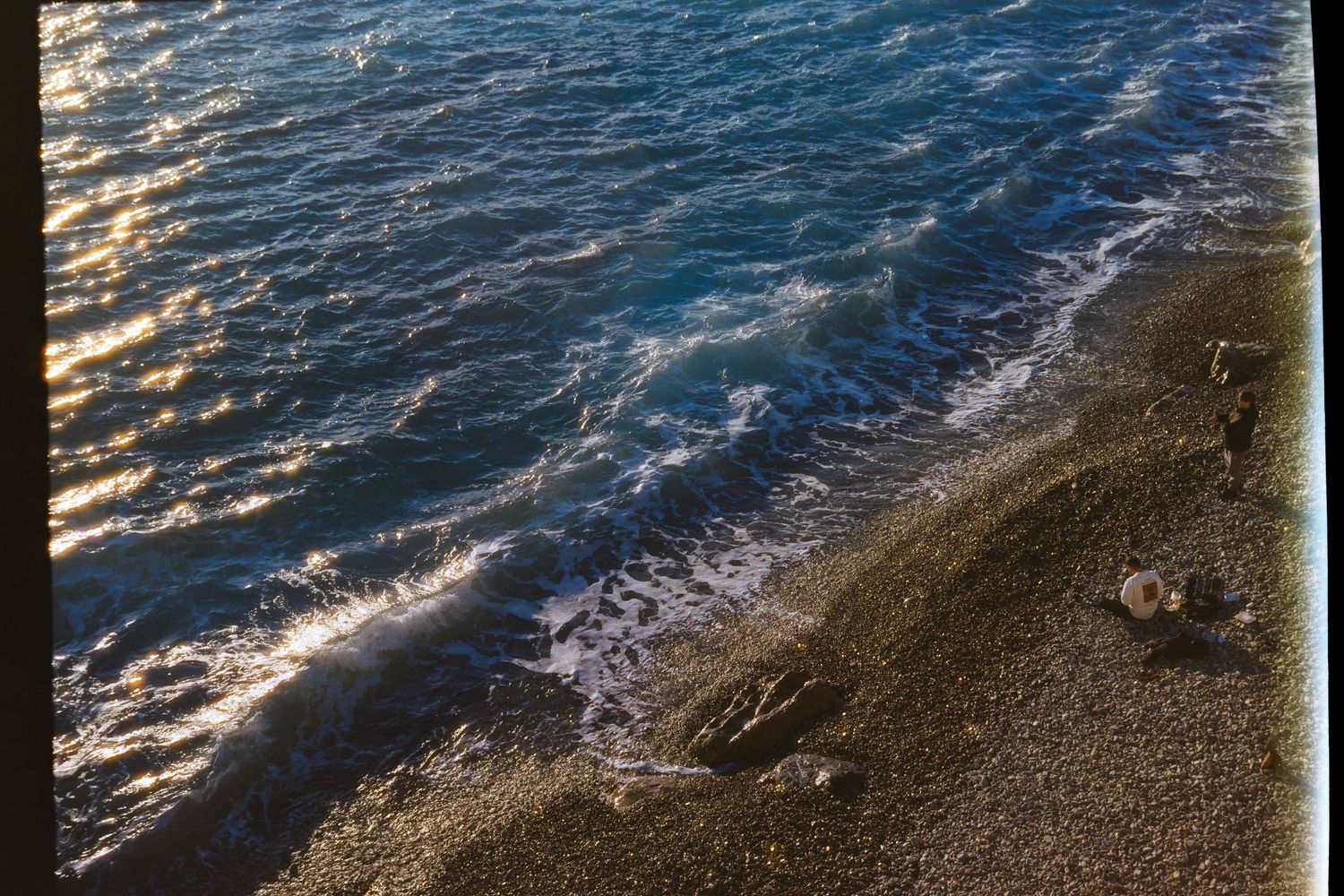 Aerial view of sunlit pebble beach with gentle waves washing ashore at golden hour. Shot on film