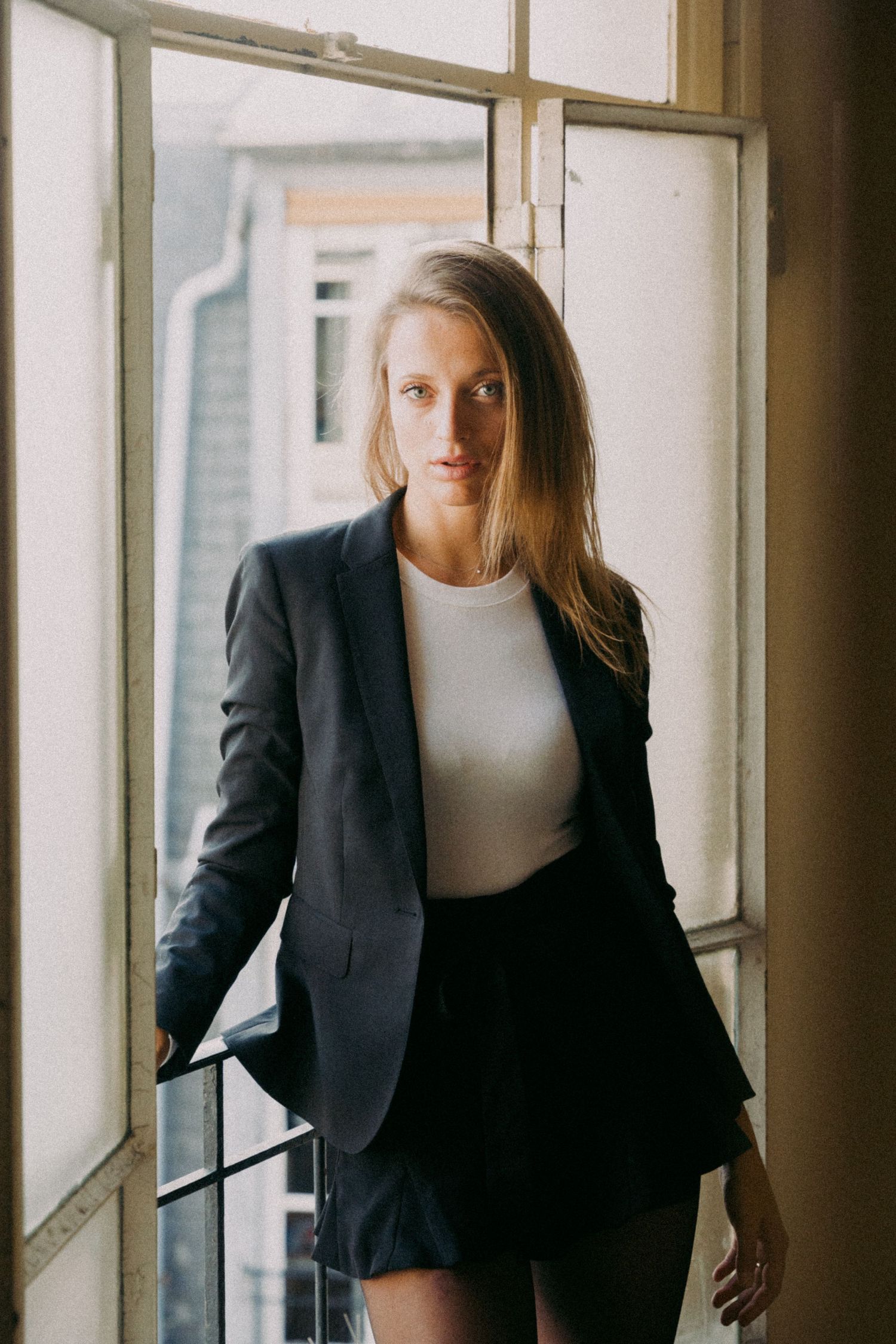 A blonde woman in a navy blazer and white top stands confidently by an open window indoors.