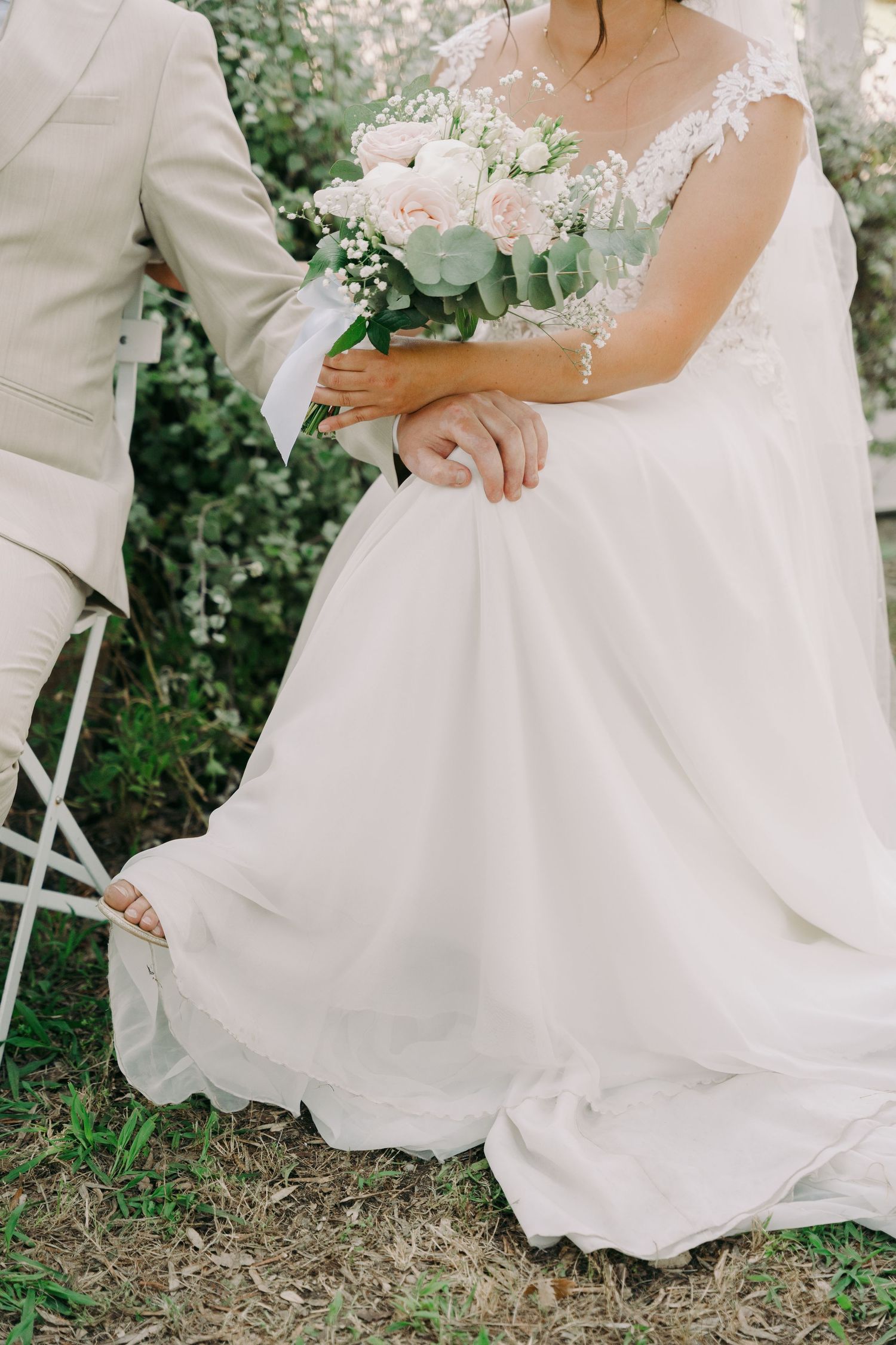 Bride in lace wedding gown holding pink bouquet beside groom in beige suit outdoors.