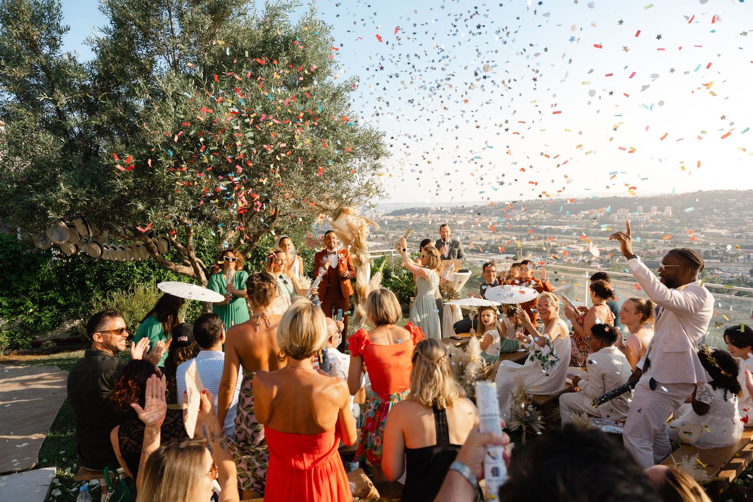 Wedding guests celebrate with colorful confetti toss at outdoor reception with scenic view.