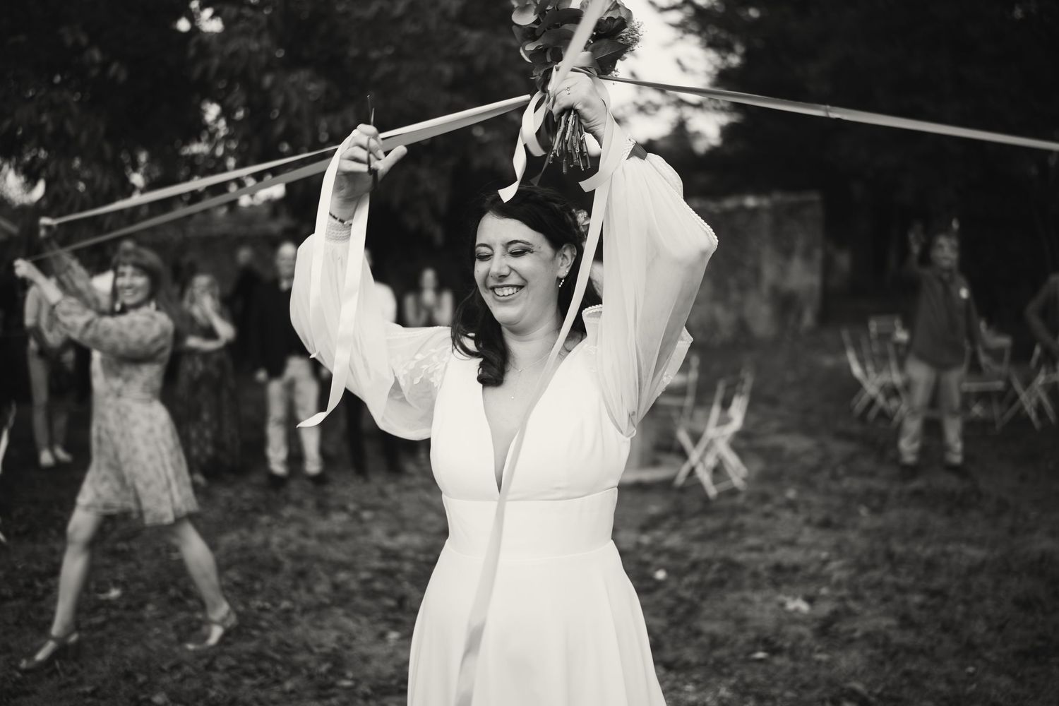 Bride in white dress laughing while hitting a pinata outdoors at a wedding celebration, black and white photo.