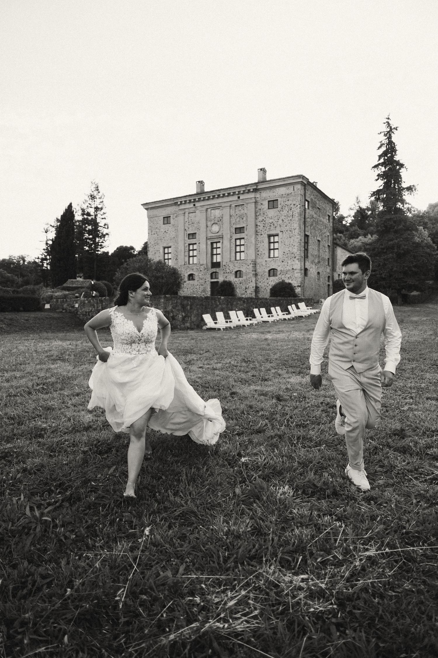Black and white photo of bride and groom running playfully in front of historic estate.