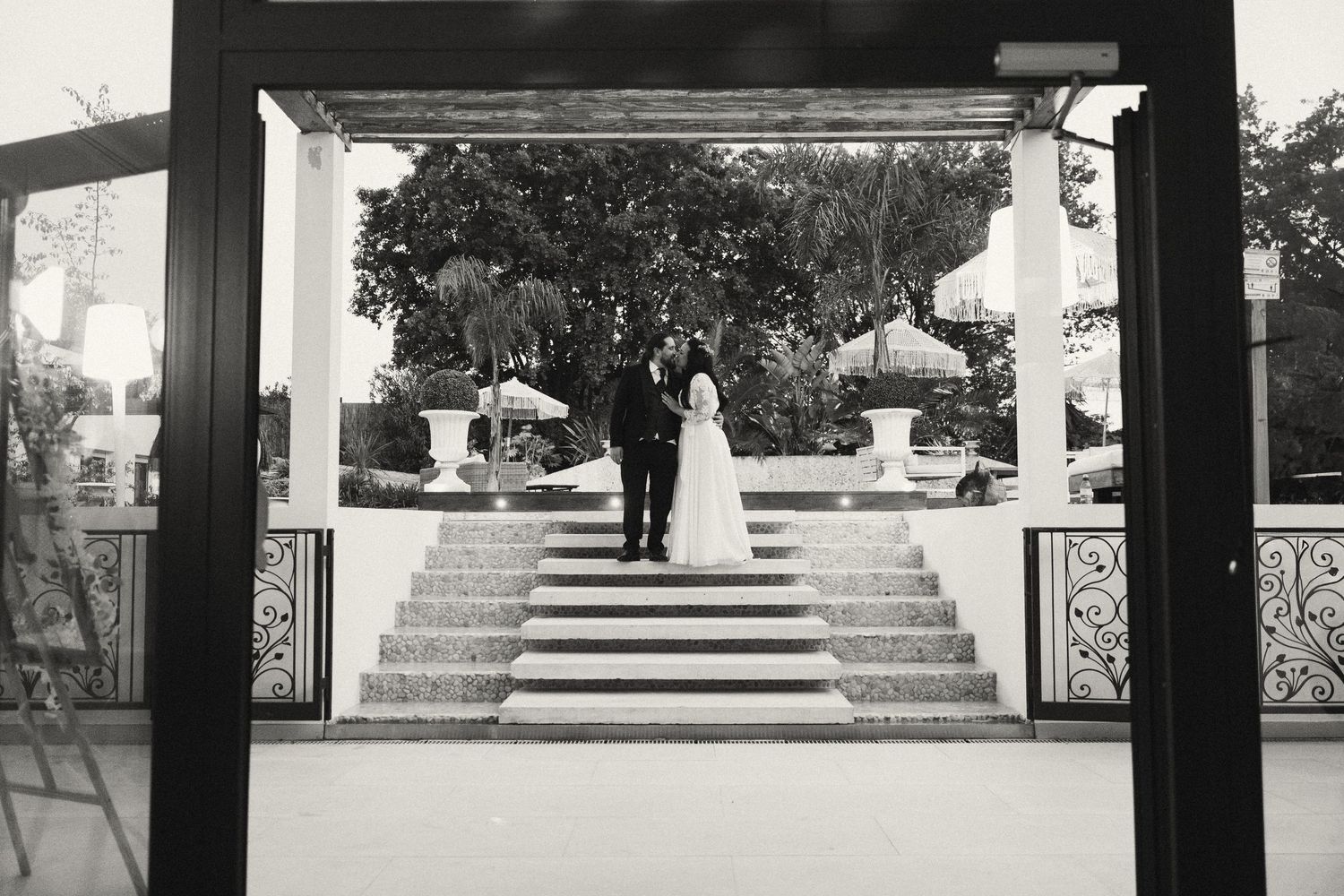 Black and white wedding photo of bride and groom standing on elegant staircase at outdoor venue.