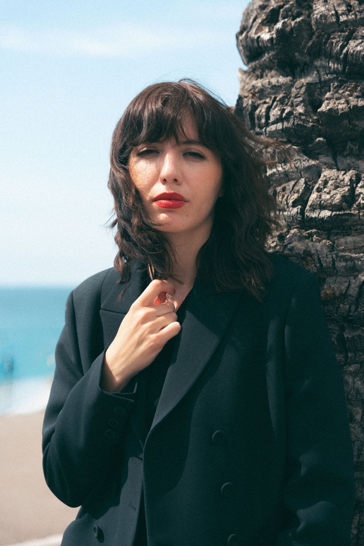 Woman with red lips in black blazer leans against textured tree trunk near the beach.
