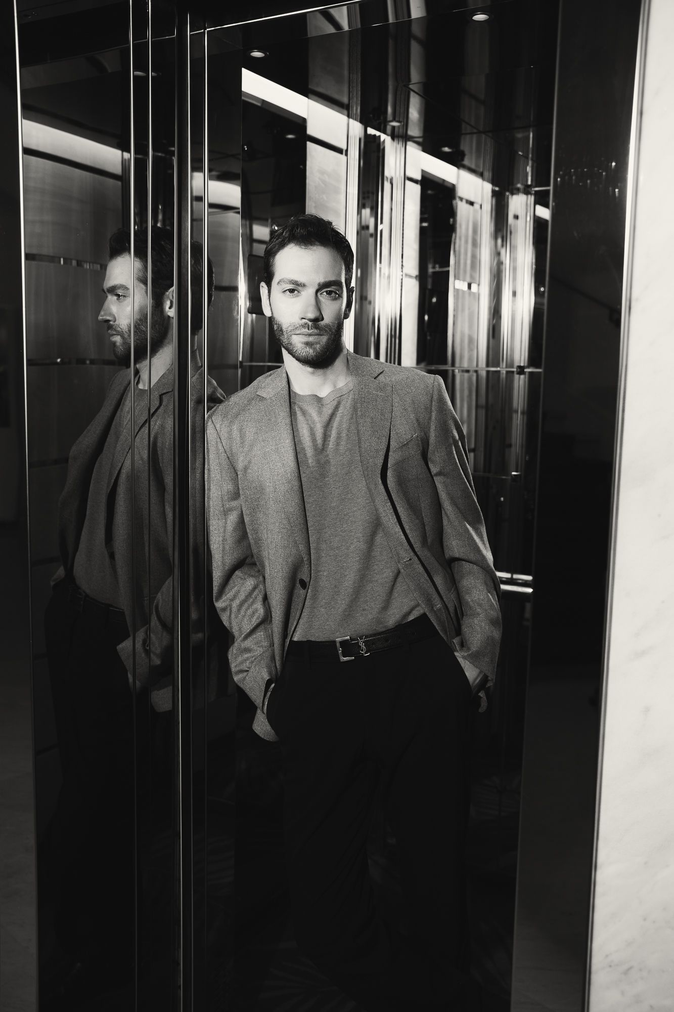 Confident bearded man in gray blazer standing in mirrored elevator in black and white.