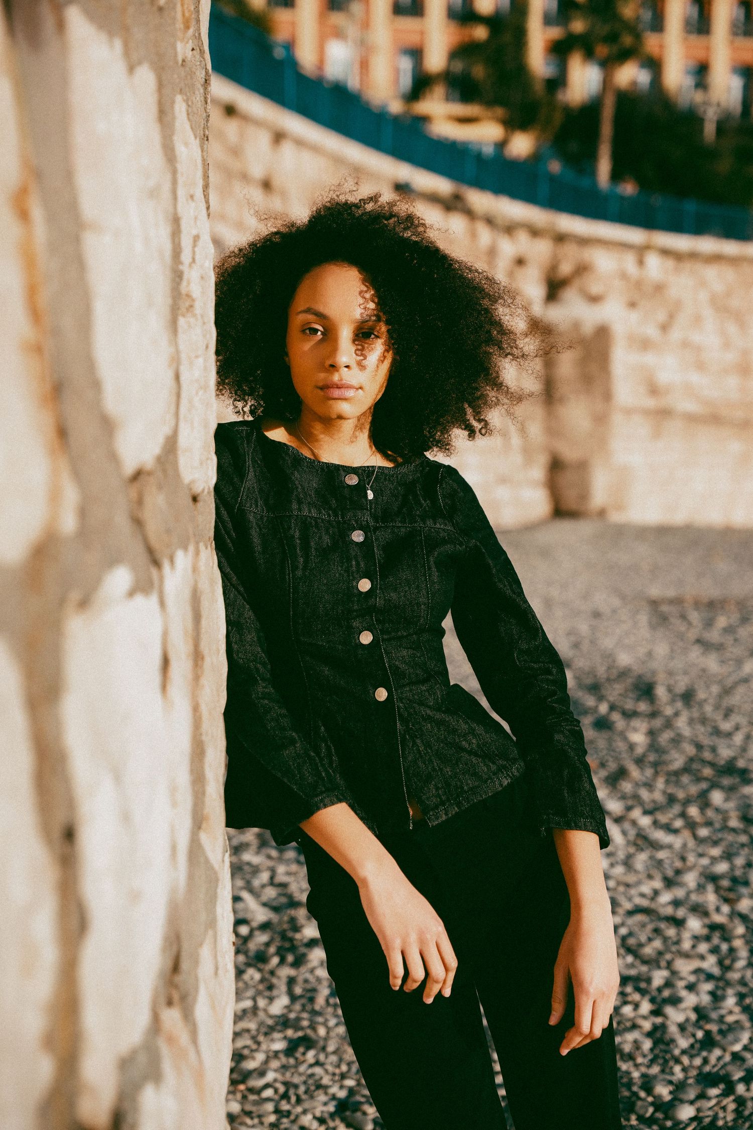 Young woman with curly hair in black velvet jacket leaning against stone wall outdoors.