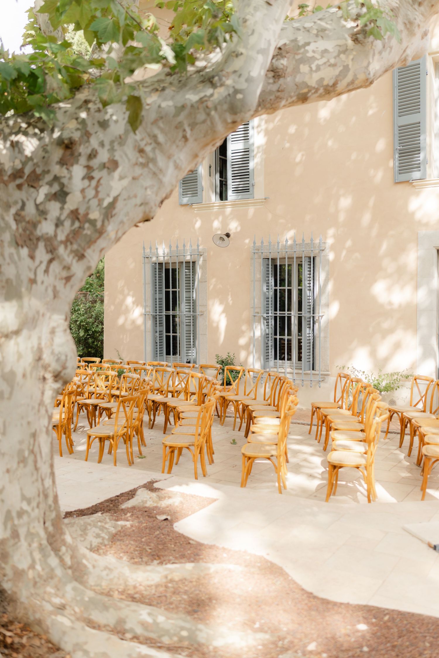 Wooden cross-back chairs arranged in rows on a sunlit terrace beside a classic French villa for an outdoor wedding ceremony.