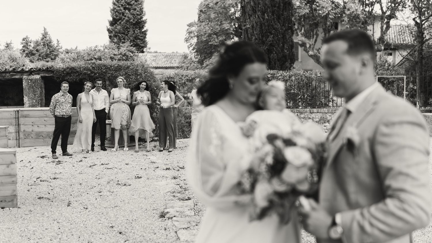 Black and white wedding photo of couple kissing in foreground while wedding guests stand together in background outdoors.
