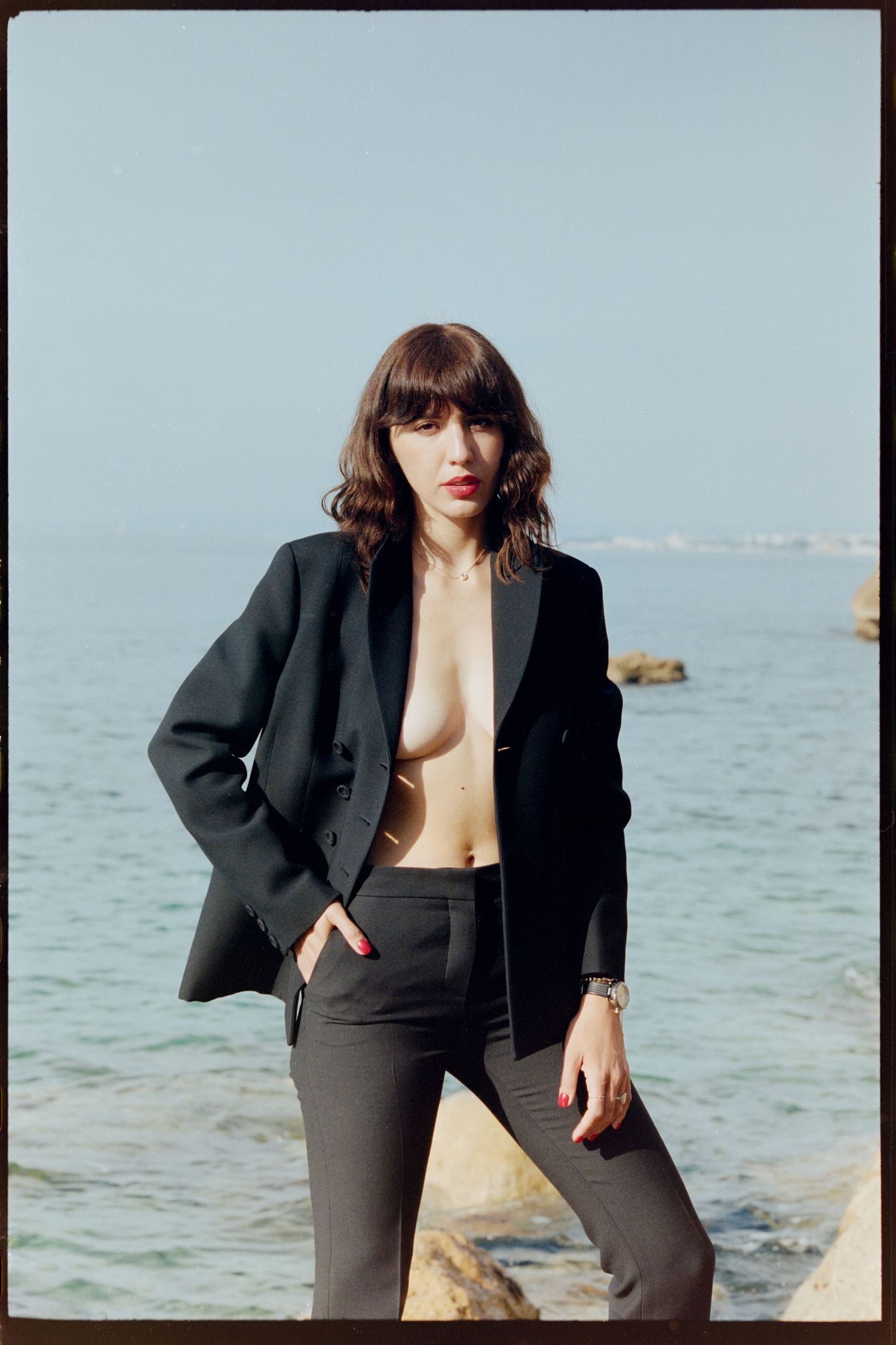 Woman in black blazer with no top poses confidently by the ocean on rocky shore.