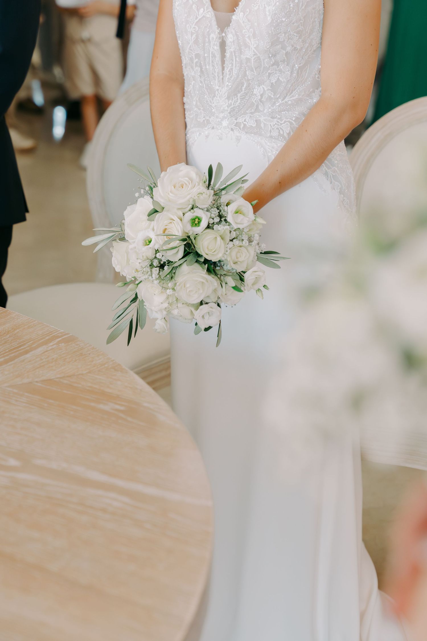 Bride in fitted white beaded gown holds elegant bouquet of white roses and greenery at wedding.