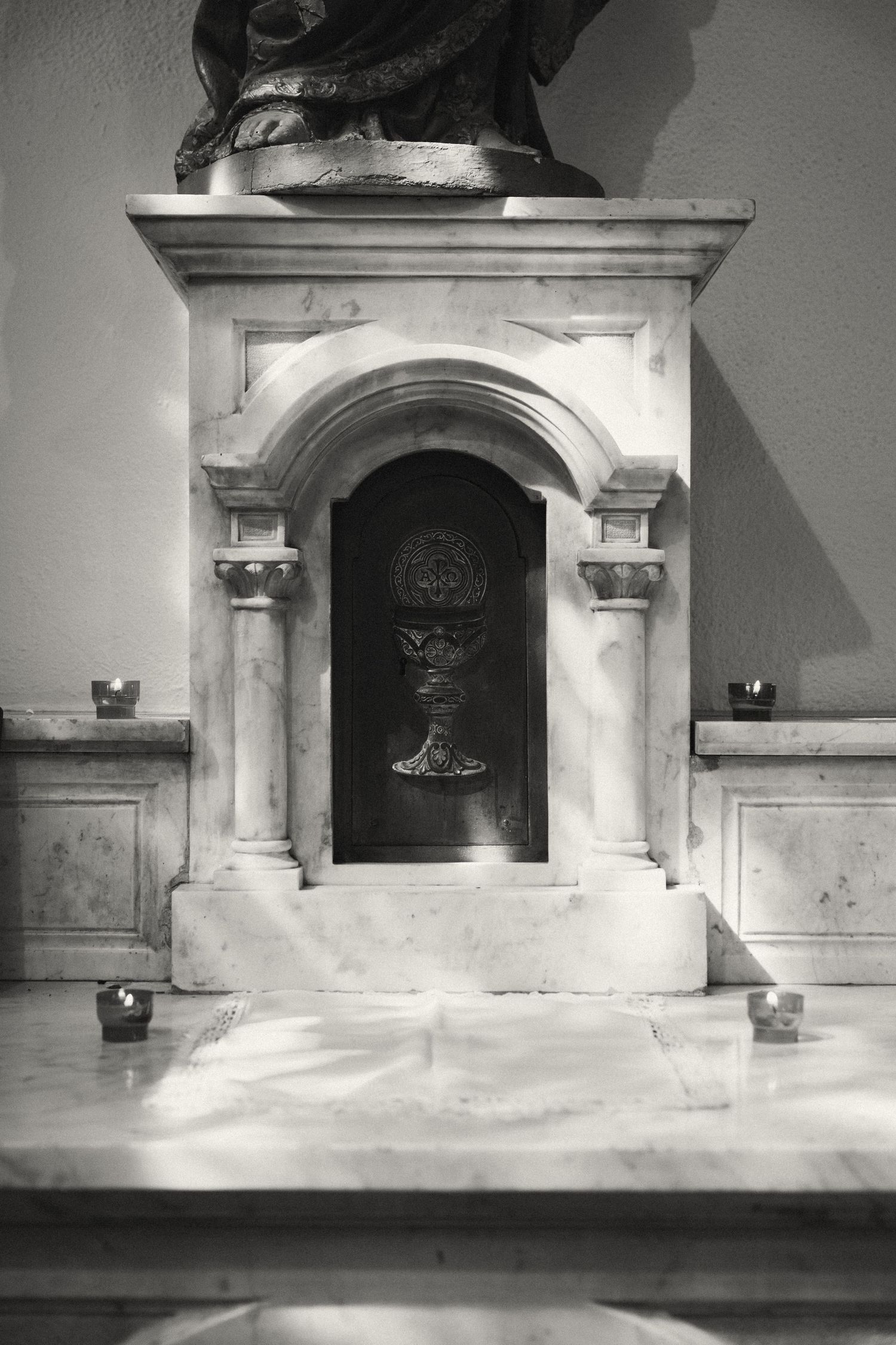 Black and white photo of a marble altar tabernacle with arched niche, flanked by candles and topped with a statue.