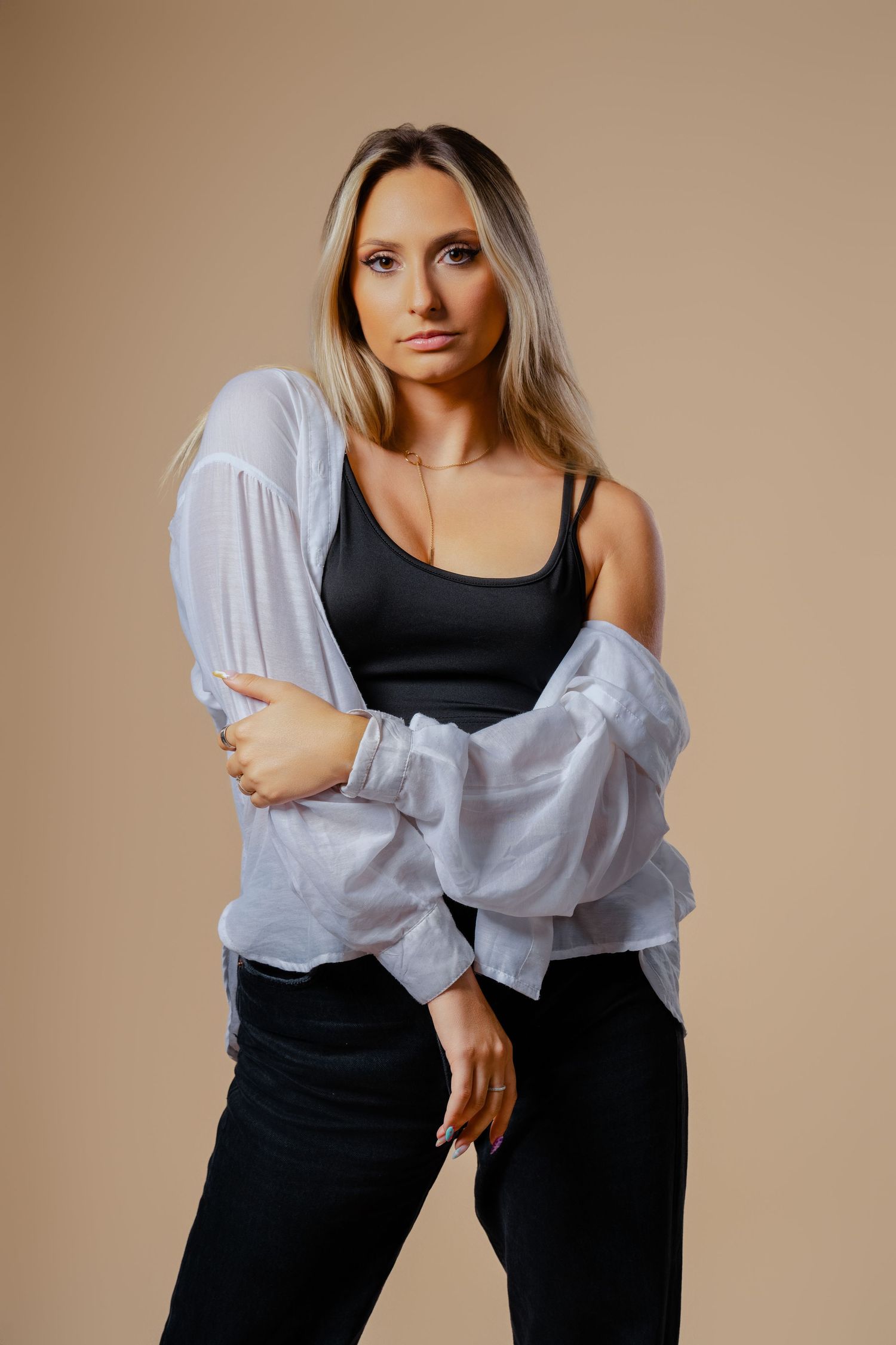 Blonde woman in black bodysuit and sheer white blouse posing against beige studio background. Dancer artist portrait