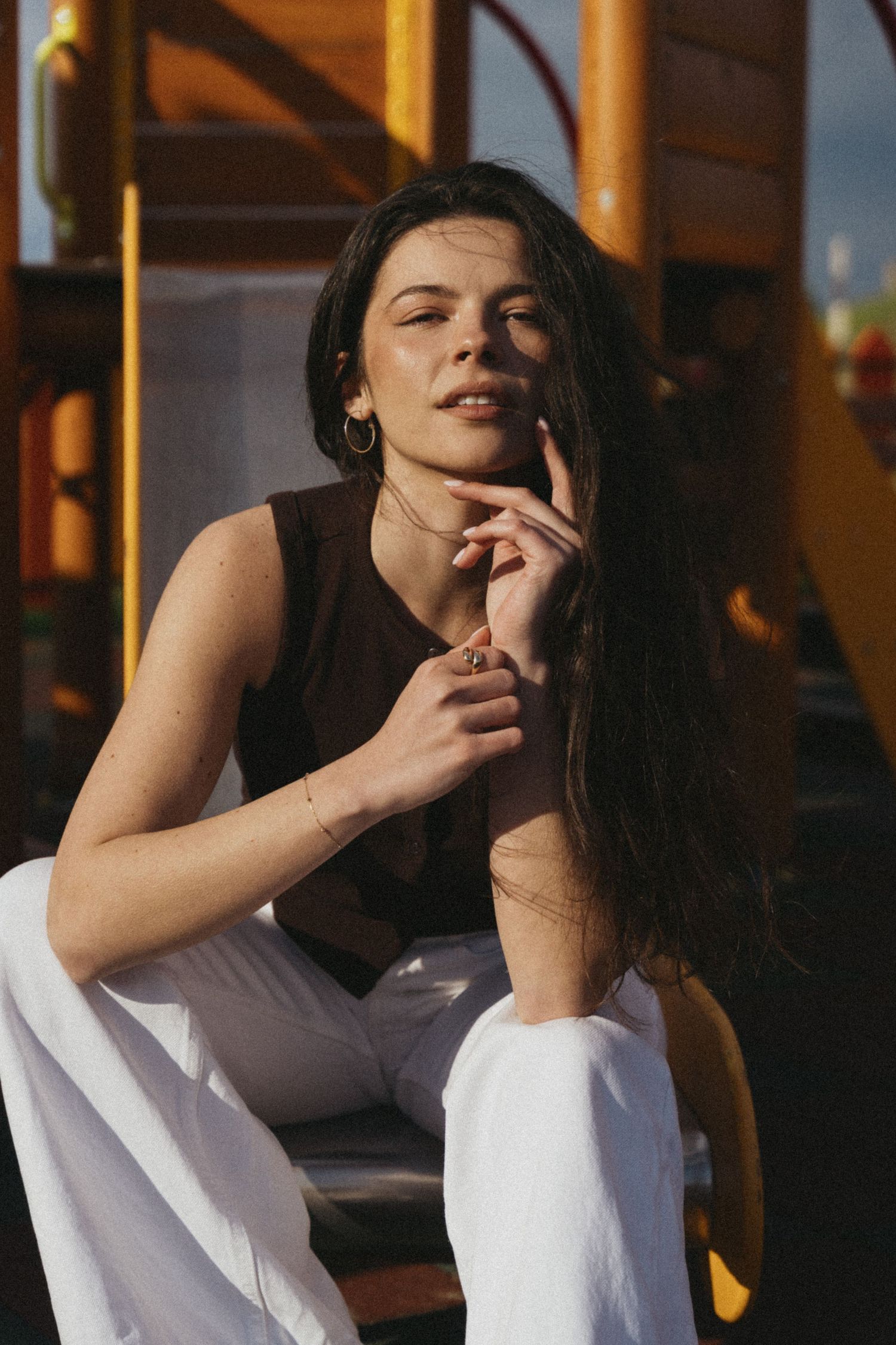 Young woman in brown top and white pants sits on playground equipment in warm golden sunlight.