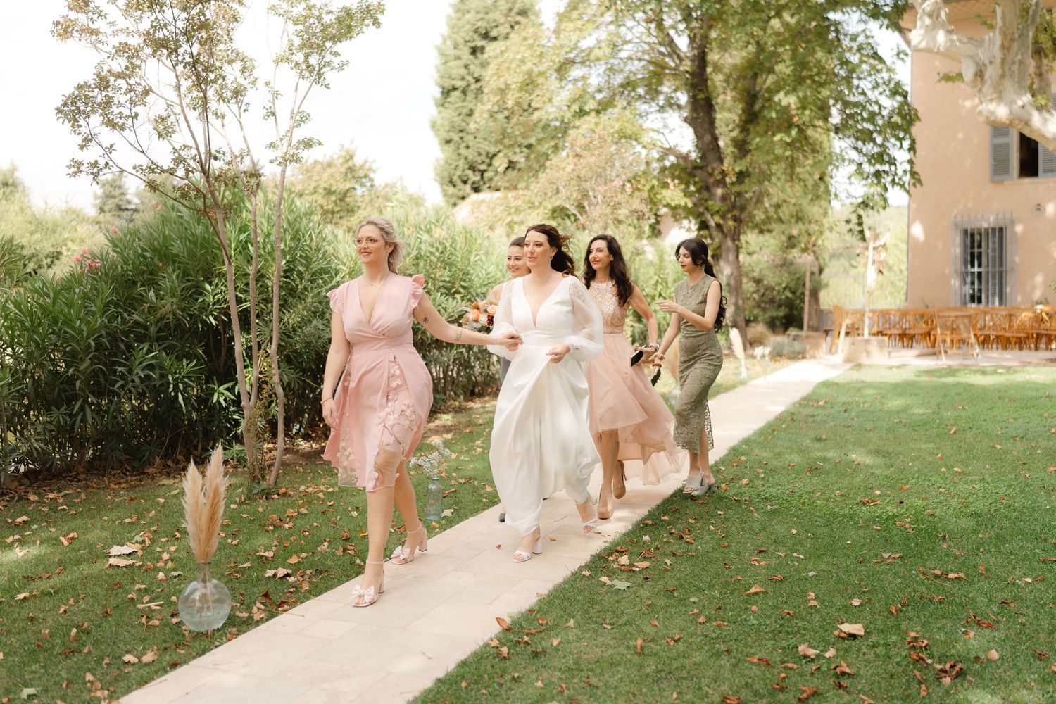 Bride and bridesmaids in pink and white dresses walking together outdoors on a garden path at a wedding.