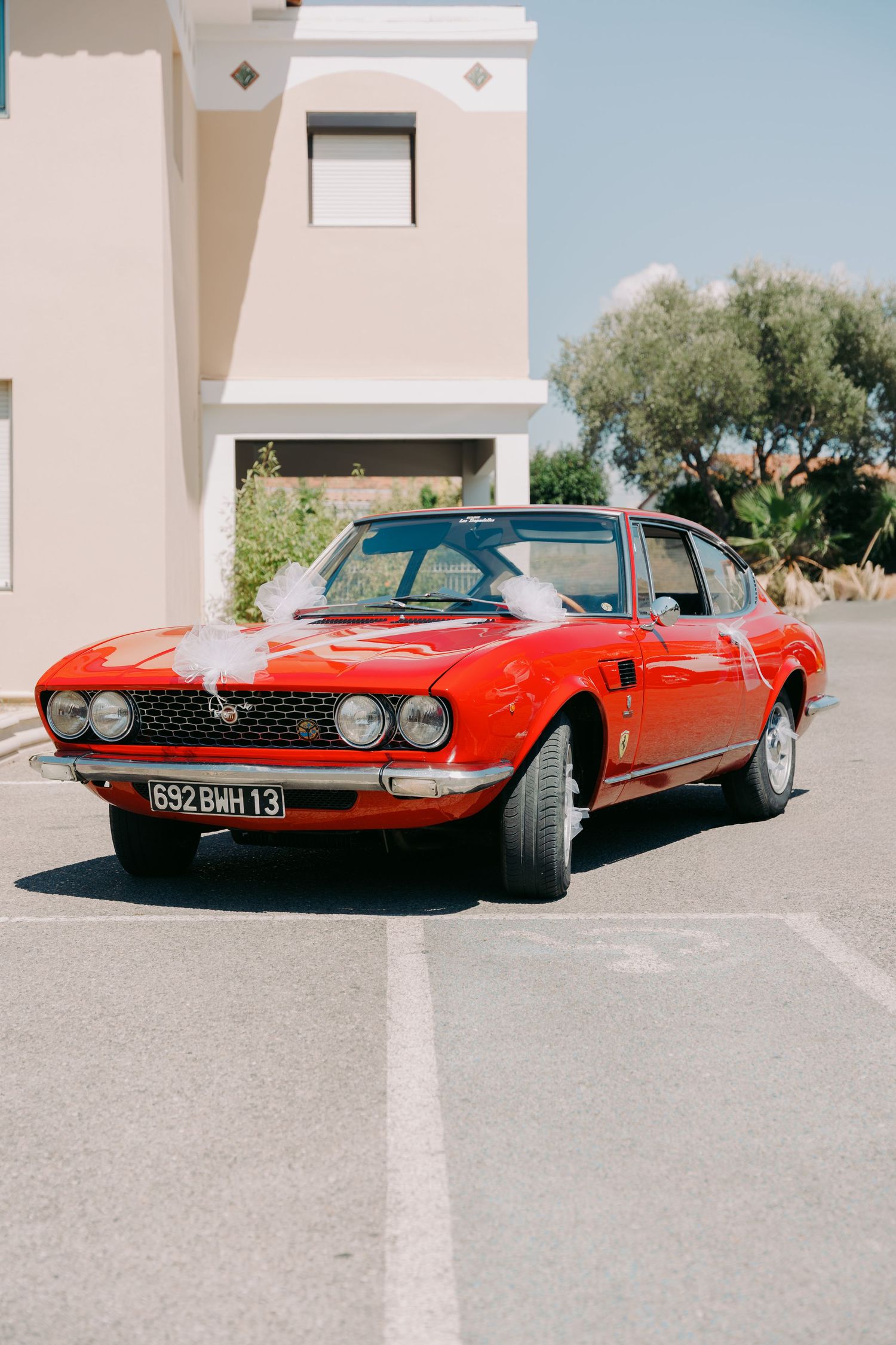 Classic red vintage Fiat sports car decorated with white ribbons parked outside on sunny wedding day.