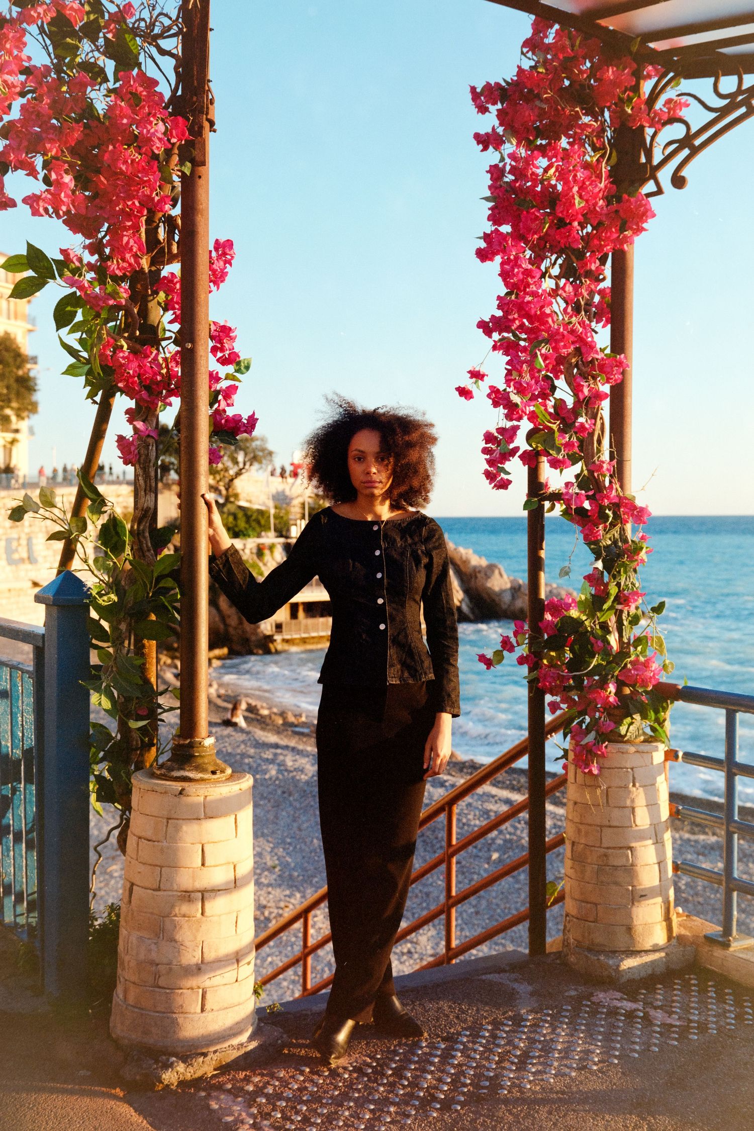 Woman in black outfit standing between floral columns with pink bougainvillea blooms overlooking a scenic ocean view.