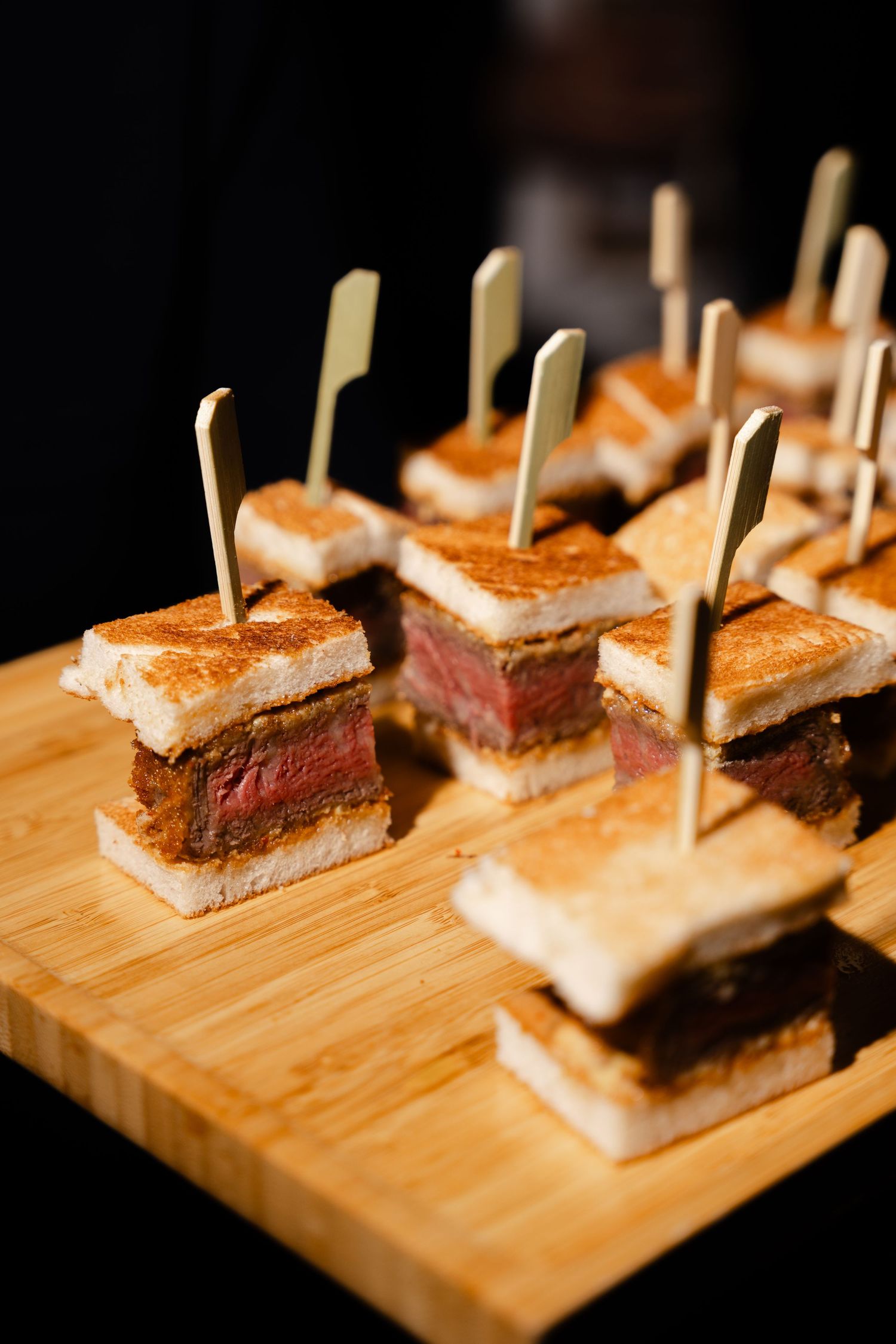 Toasted steak finger sandwiches with toothpicks arranged on a wooden serving board.