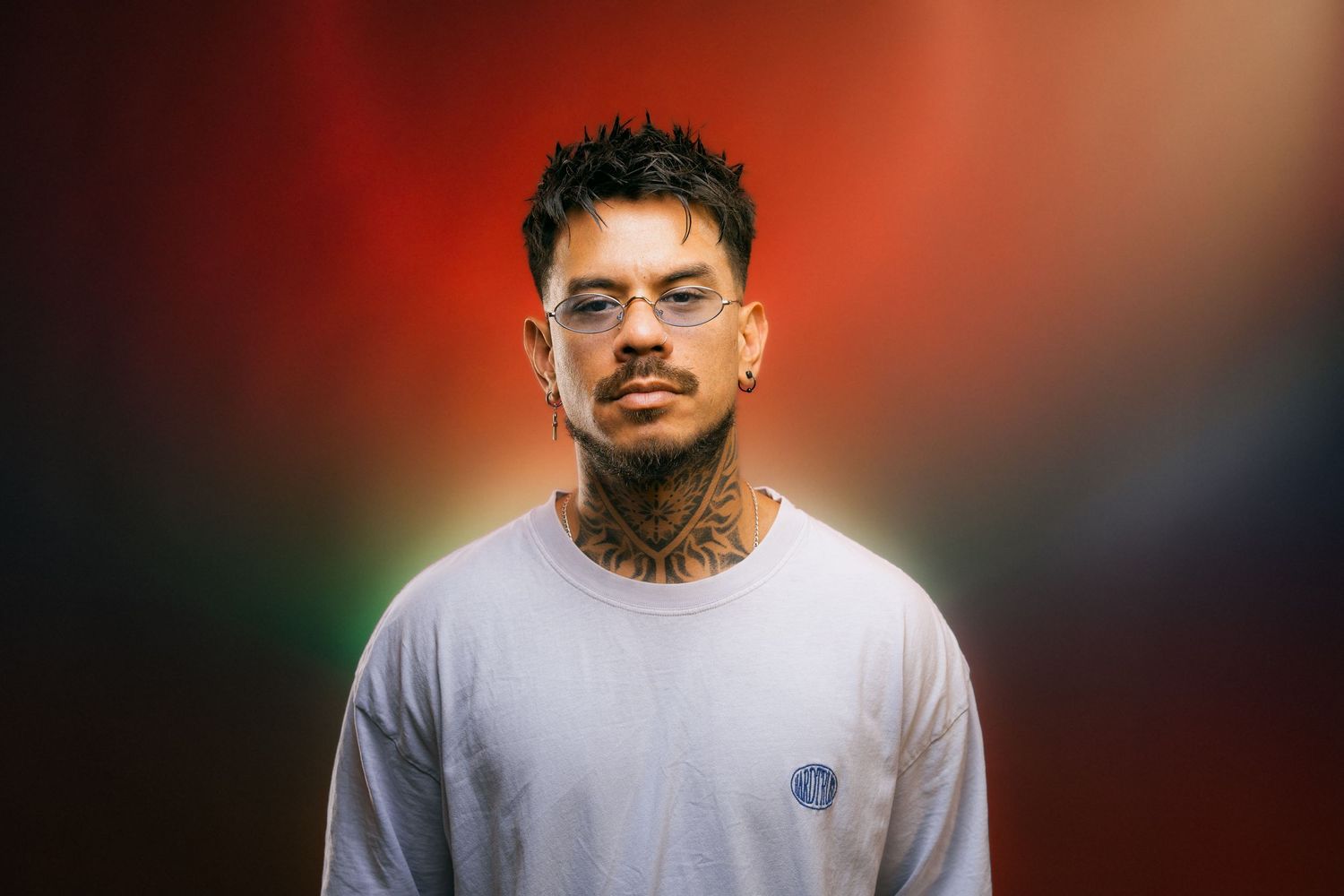Young man with neck tattoos in a white t-shirt posing against a colorful red and green gradient background. DJ portrait