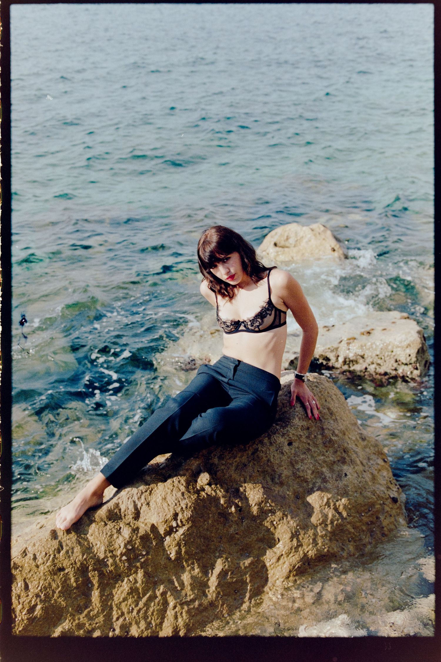 Woman in lace bralette and black pants poses on coastal rock with ocean behind her.
