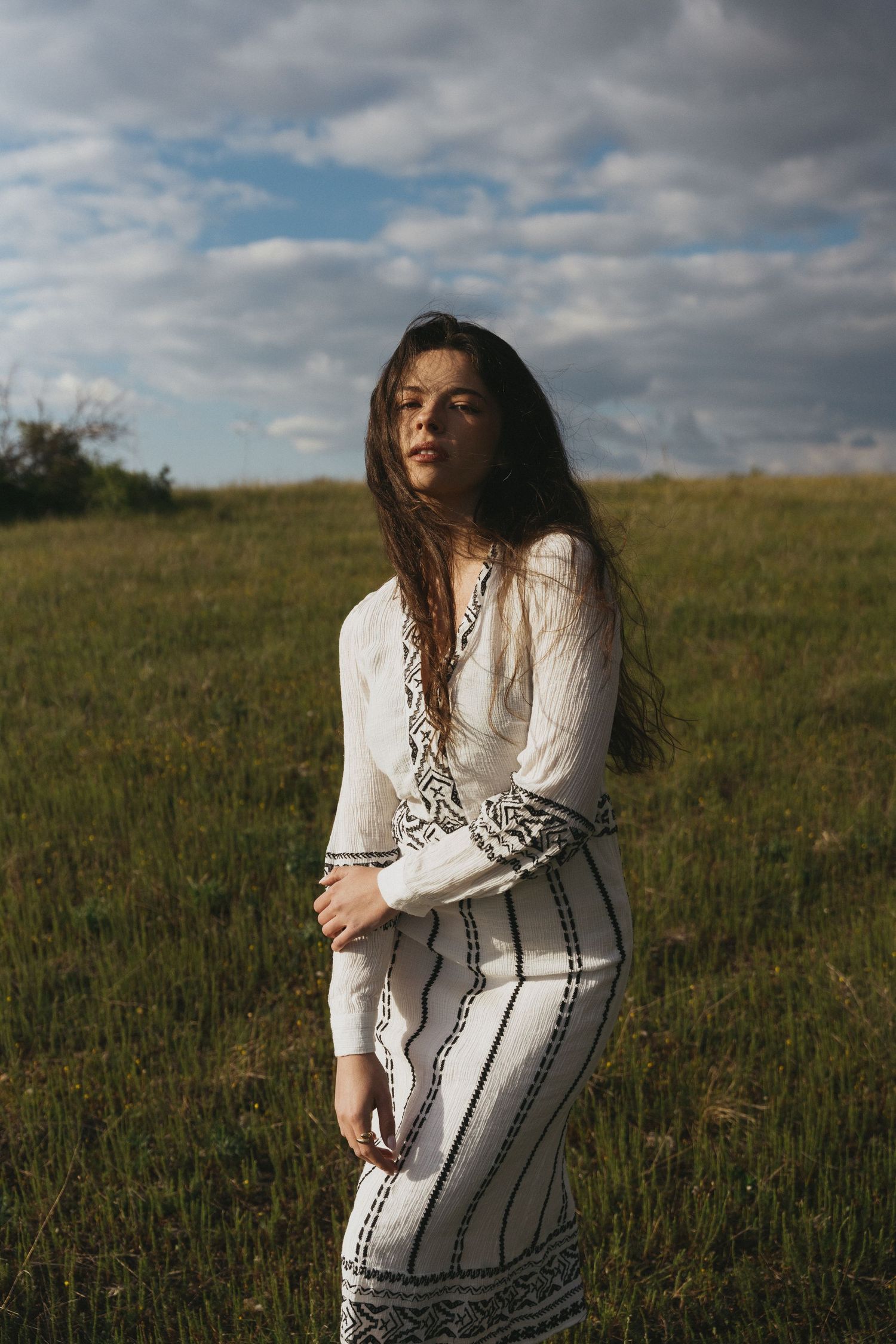 Young woman in white embroidered boho dress stands in green field under cloudy sky.