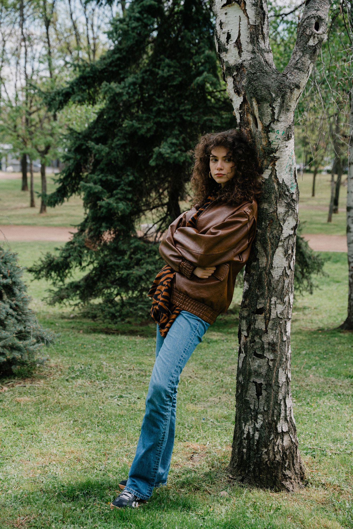 Full-length shot of curly-haired woman in brown leather jacket and flared jeans by birch tree.