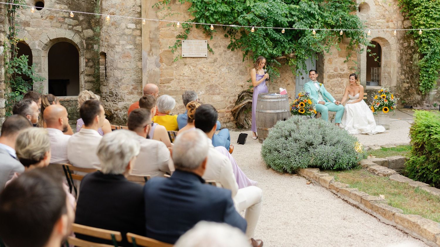 Outdoor wedding ceremony in a rustic stone courtyard with guests seated on wooden chairs facing the couple.
