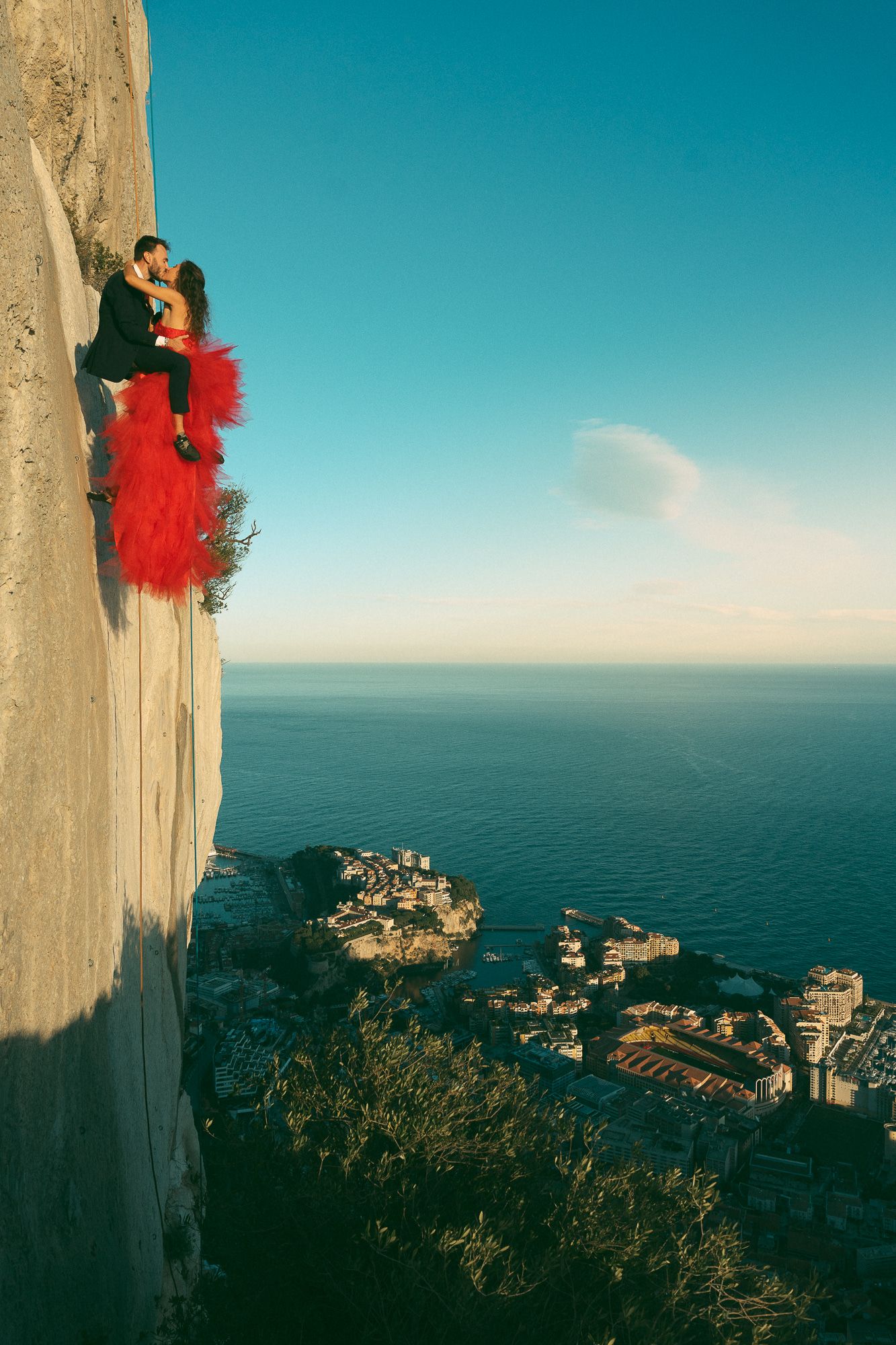 Couple in formal wear kissing on a cliff edge overlooking a coastal town and ocean at sunset.