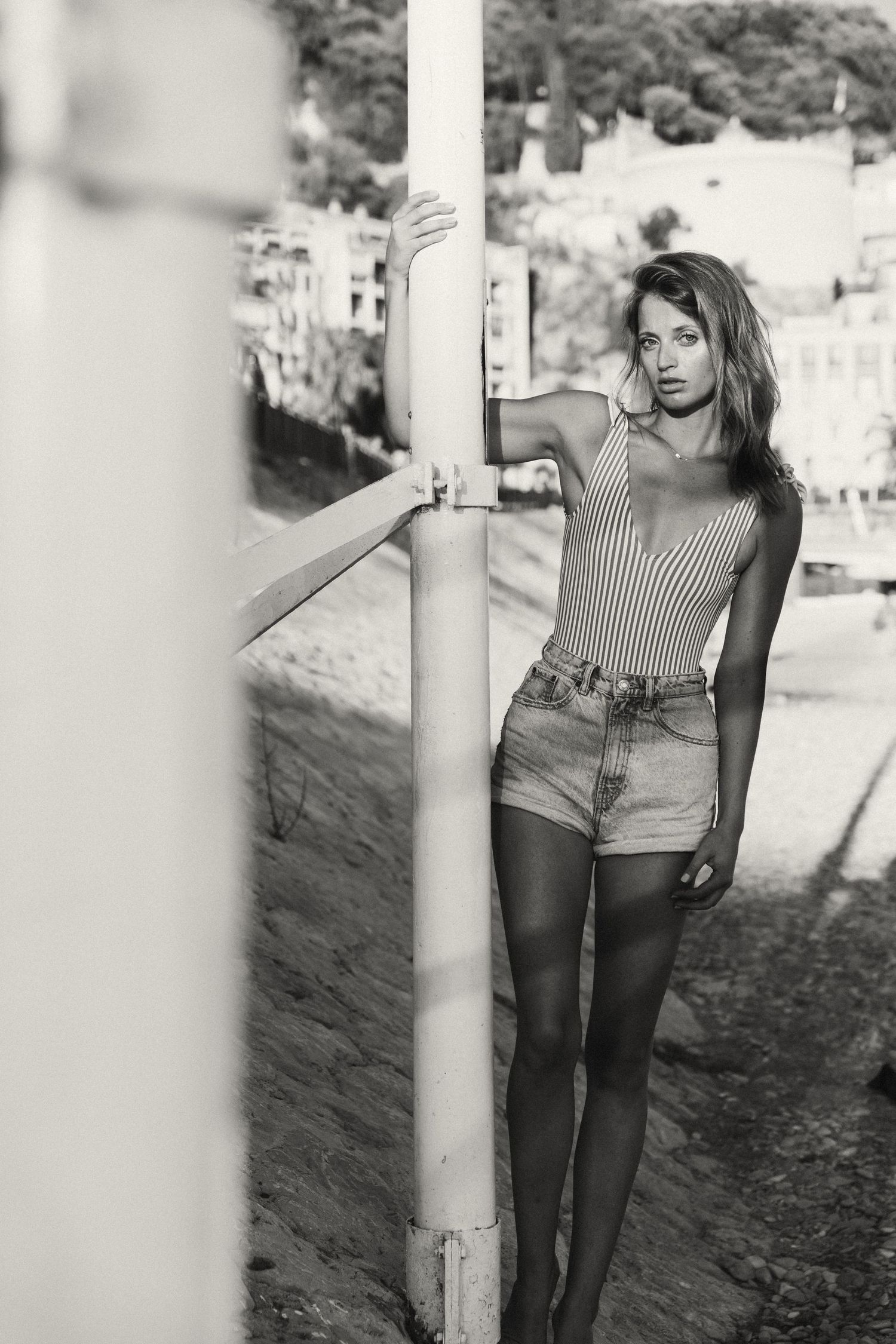 Black and white photo of young woman in striped bodysuit and denim shorts posing by metal pole.
