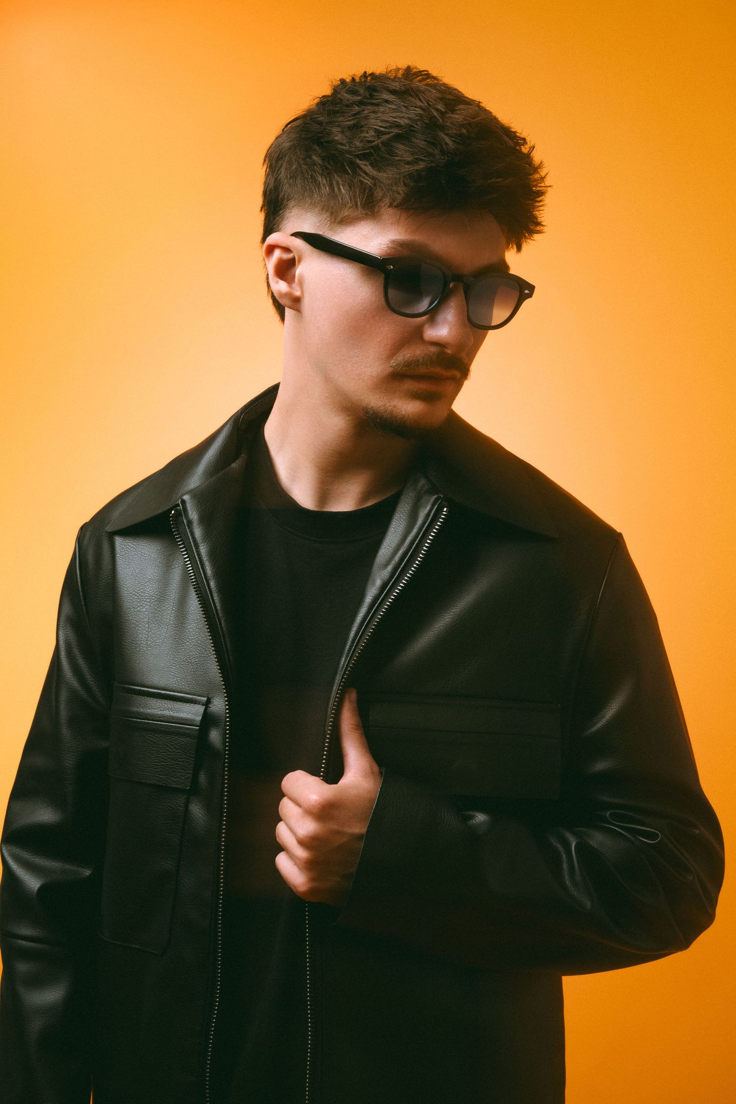 Young man in a black leather jacket and glasses posing against a vibrant orange studio background.