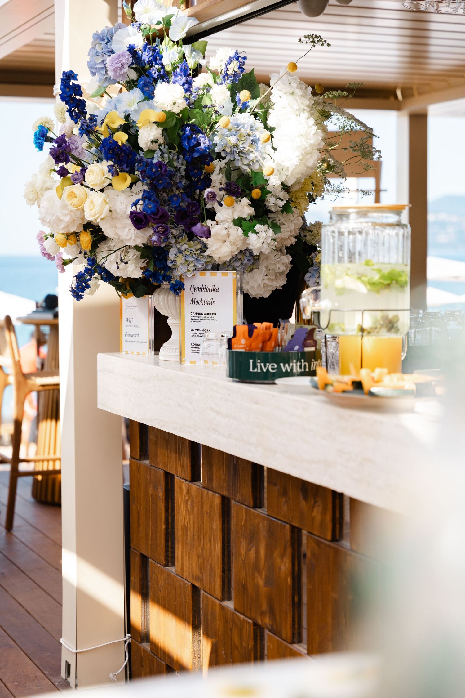 Large blue and white floral arrangement on a wooden bar counter with juice dispensers and snacks.