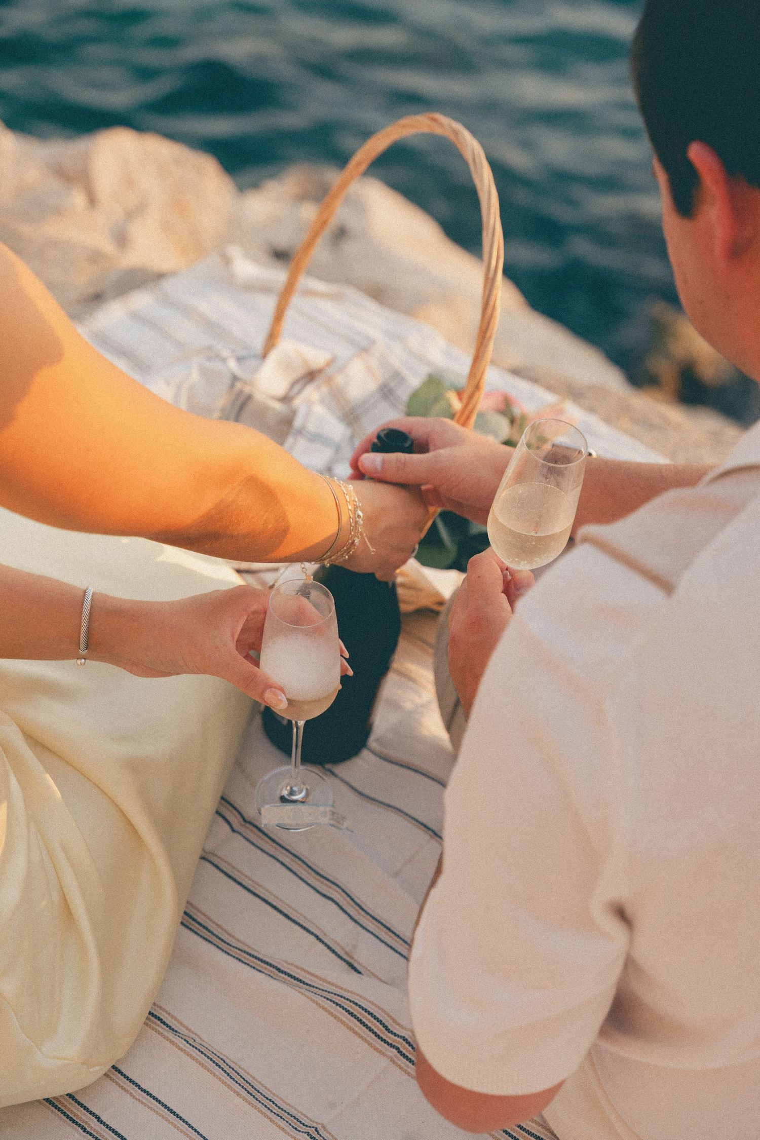 Couple sharing champagne glasses on picnic blanket by the sea during golden sunset.