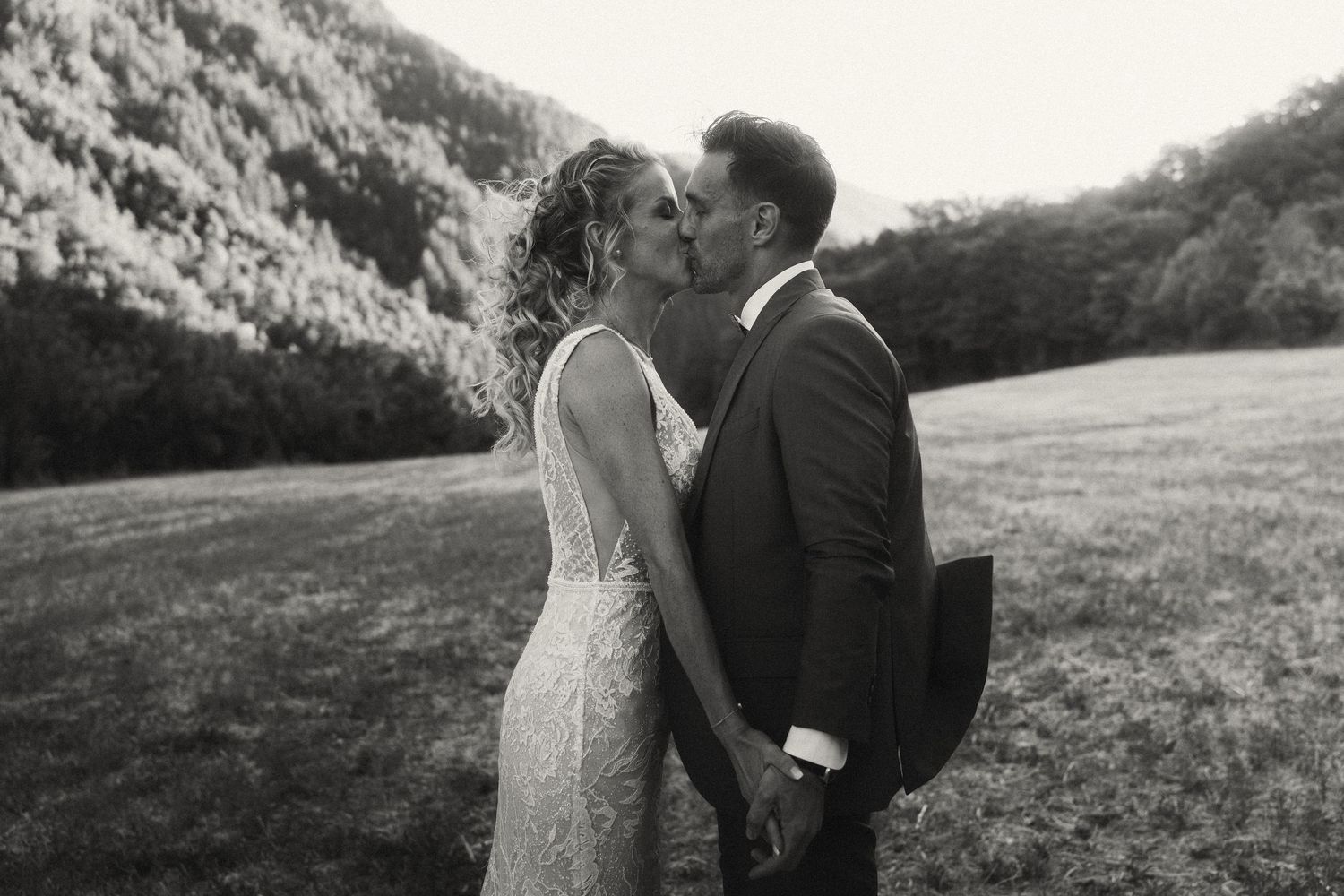 Black and white wedding photo of bride and groom sharing a romantic kiss in a scenic outdoor meadow.