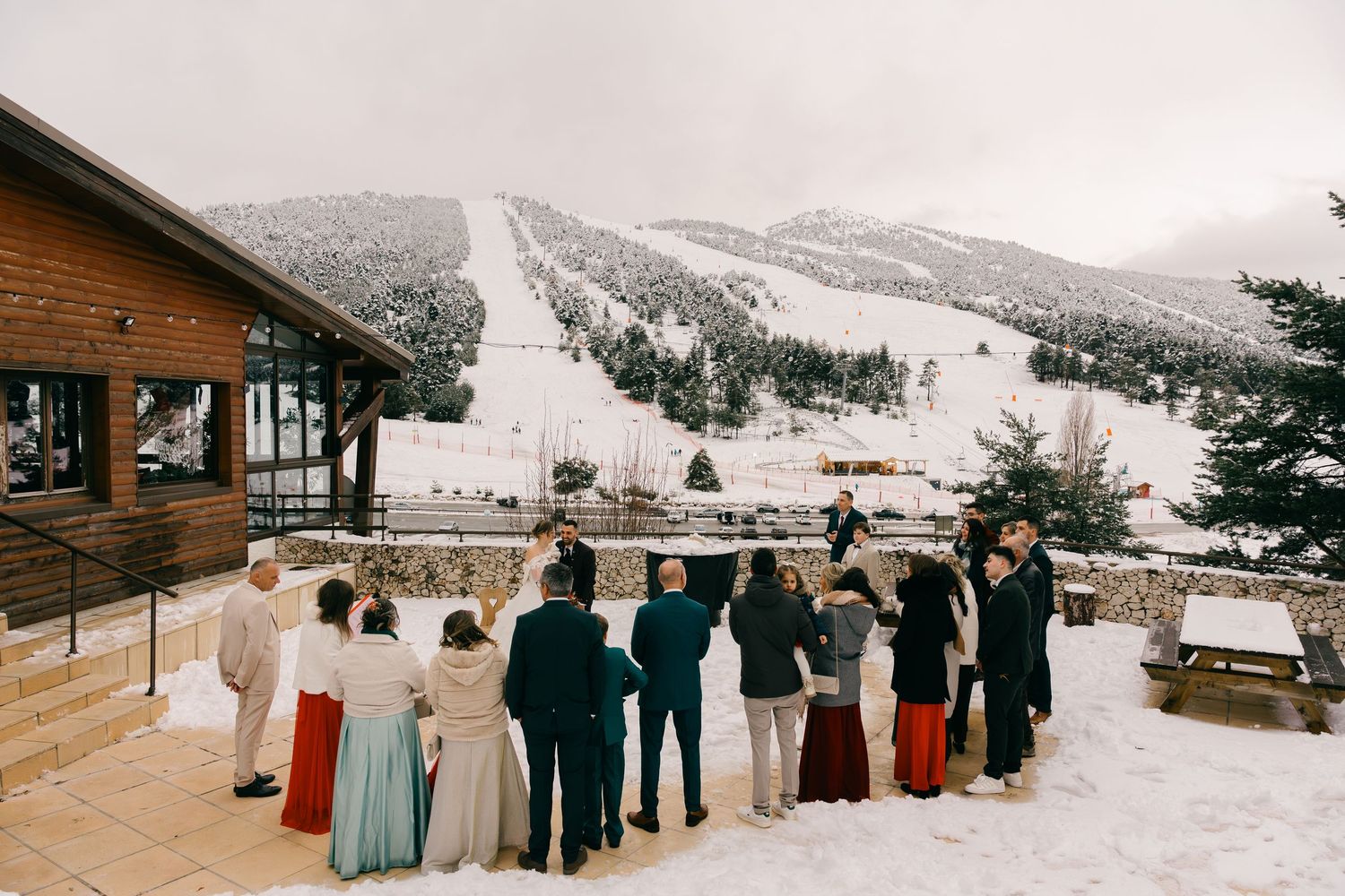 Wedding guests gather outdoors on a snowy terrace with a ski slope backdrop in winter.