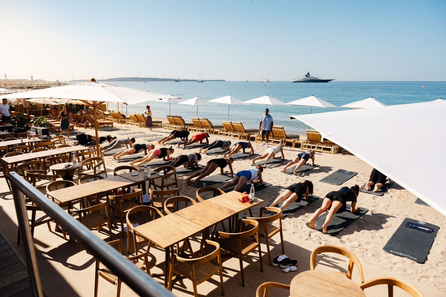 Group yoga class on a sunny beachside terrace with ocean and yacht views in background.