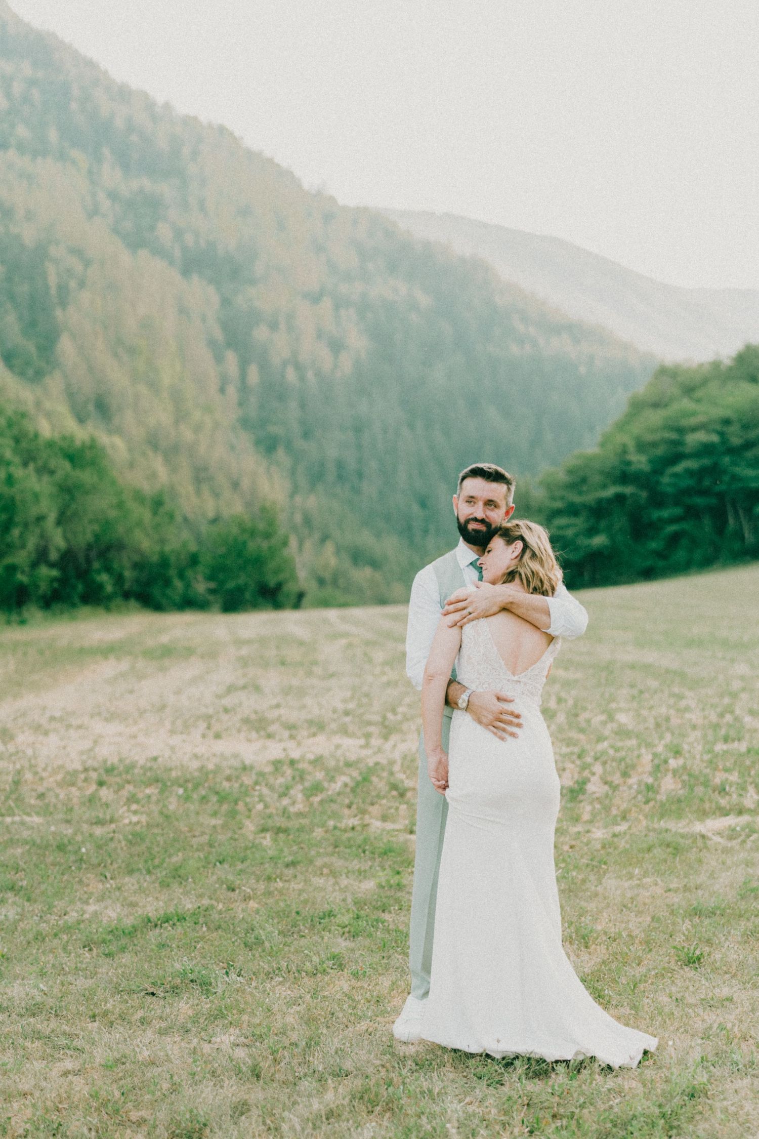 Bride and groom embrace in a scenic mountain meadow, bride in white gown, surrounded by lush green hills.