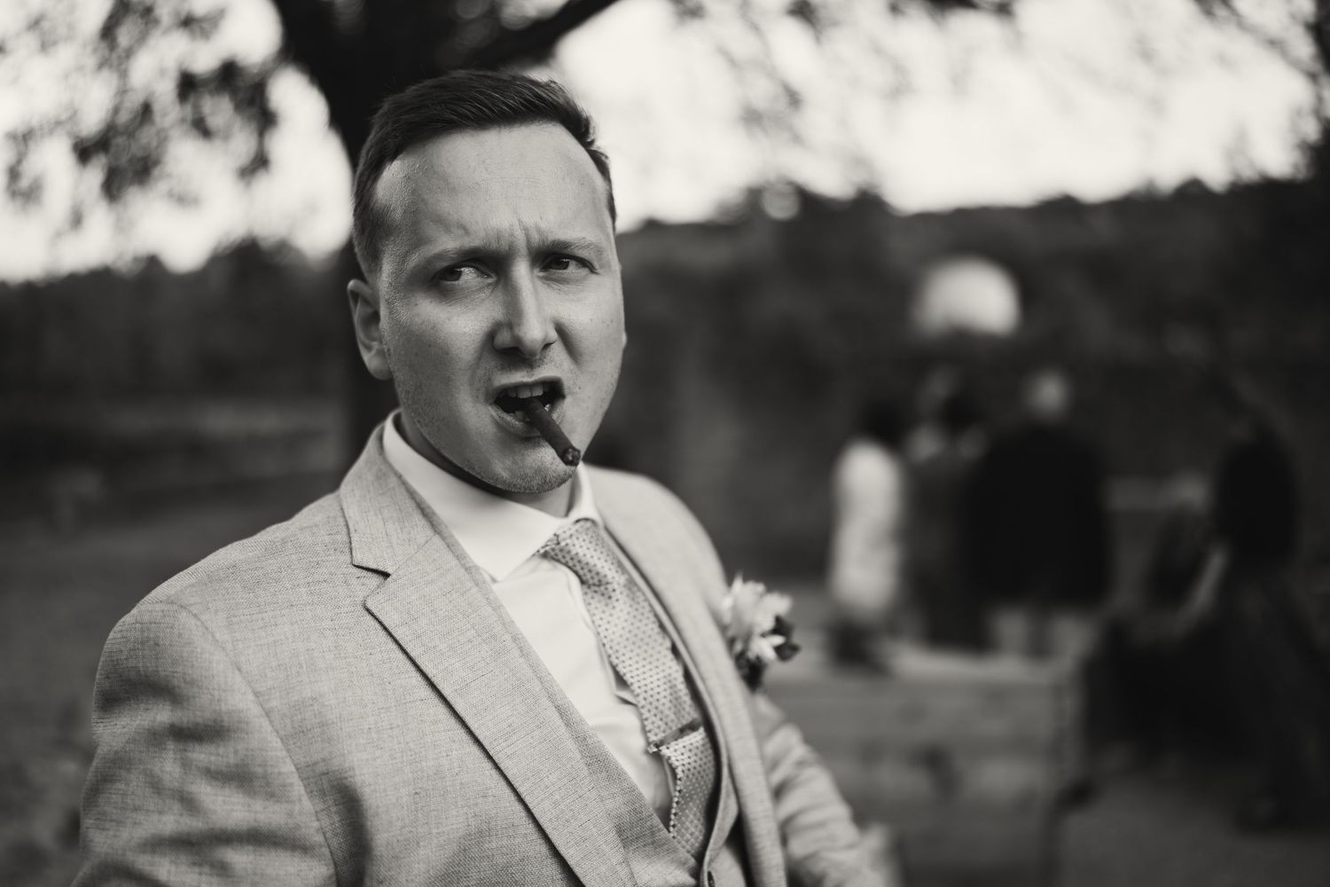 Black and white photo of a groom in a suit with boutonniere, holding something in his mouth at an outdoor wedding.