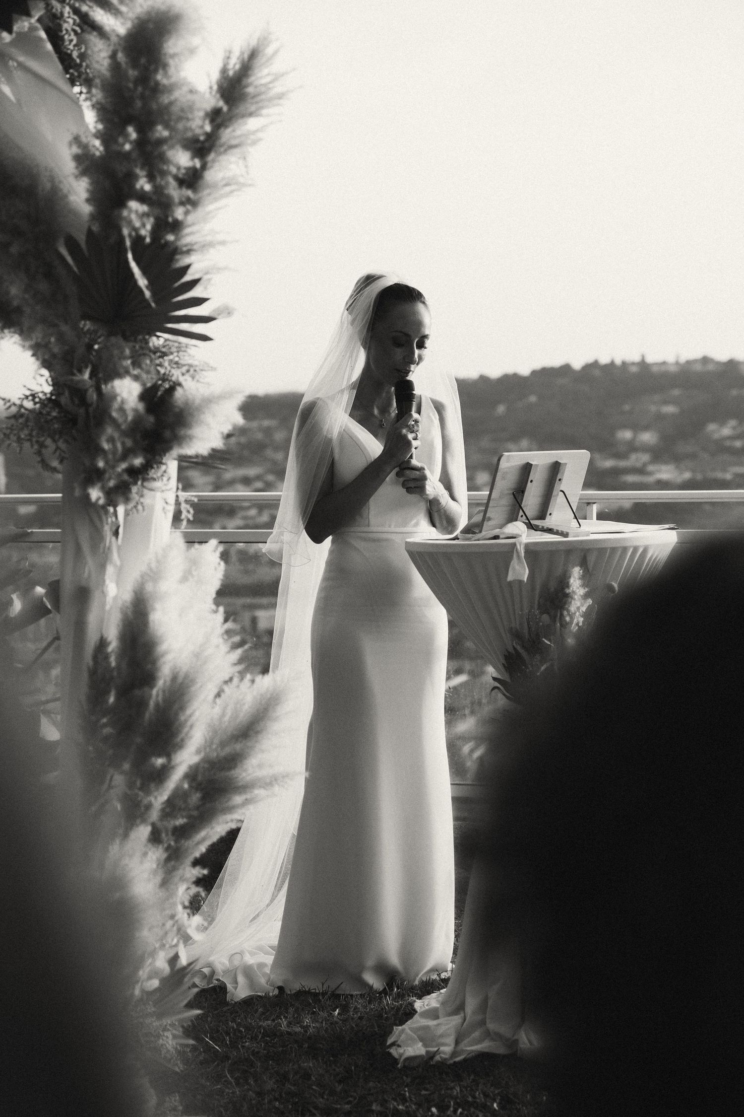 Bride in white gown and veil speaks at outdoor wedding ceremony in black and white photo.